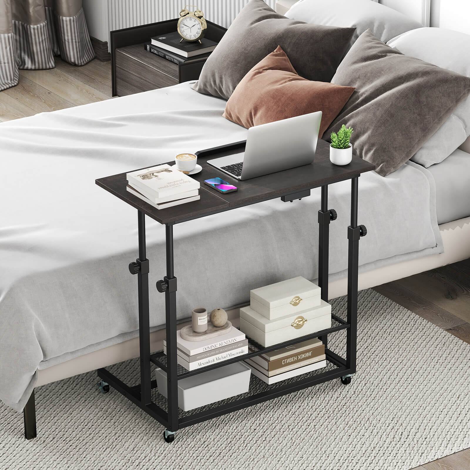 The image features a bed with a laptop on a bedside table. The table also has a clock, a cup, and a book on it. There are two pillows on the bed, one on the left side and another on the right side. The bed is covered with a white sheet, and there is a rug placed underneath it. The scene appears to be a cozy and comfortable space for work or relaxation.