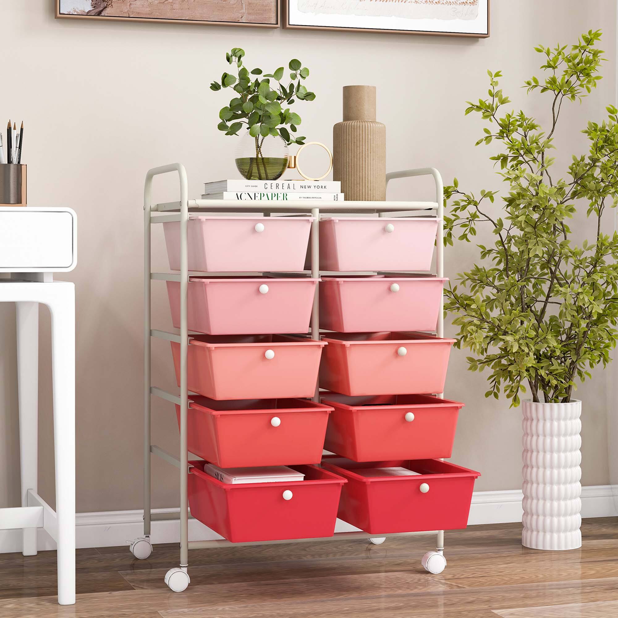 The image features a red cart with a shelf and several pink drawers. The cart is placed on a wooden floor, and there are two potted plants nearby. The cart also has a book and a vase on it. The scene is set in a room with a white wall, and there is a table in the background.