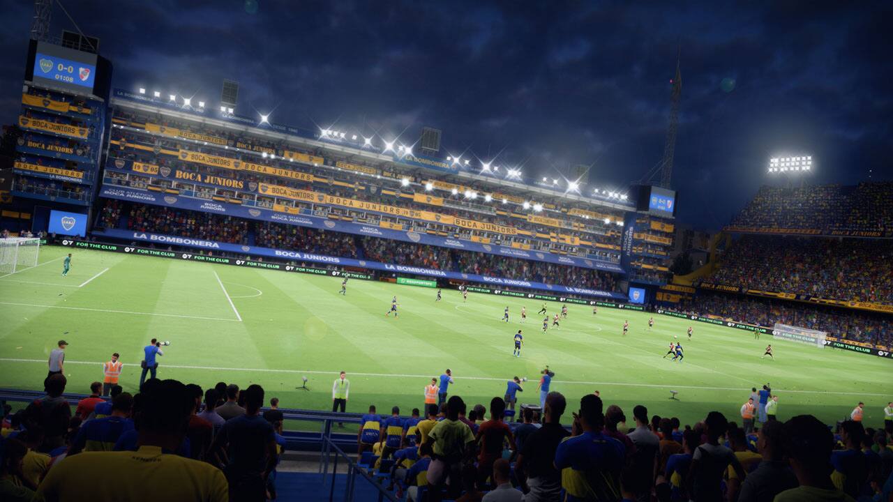 The image shows a soccer game in progress, with a large crowd of spectators watching the match. The text on the image reads "Boca Juniors," indicating that the game is taking place in a stadium belonging to the Boca Juniors soccer team. The game is currently at a 0-0 tie, with the clock showing 1:08. The spectators are spread out around the field, with some sitting on benches and others standing. The atmosphere appears to be lively and engaging, as the crowd eagerly watches the players on the field.