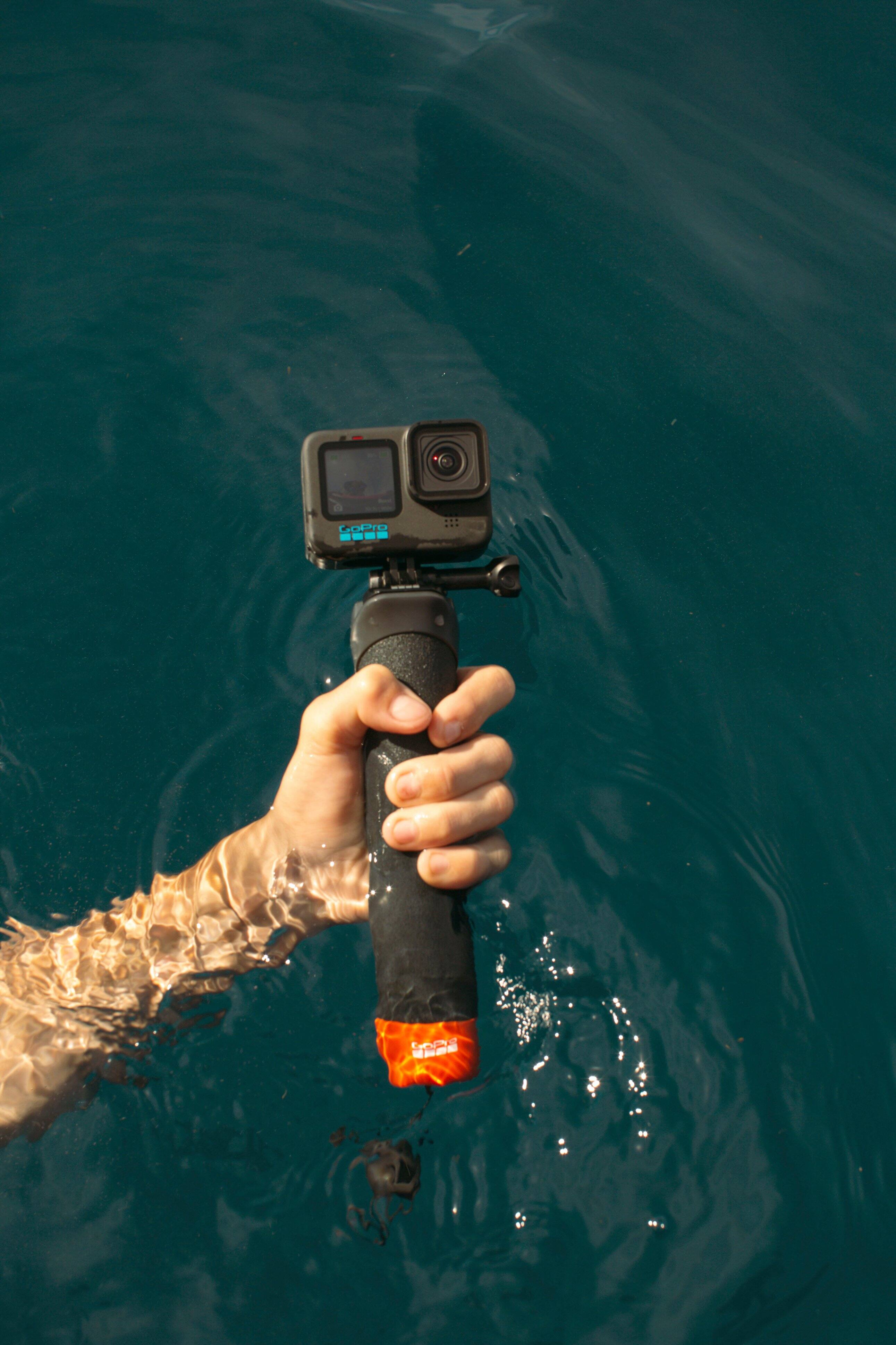 The image shows a person holding a GoPro camera while swimming in the ocean. The camera is attached to a pole, which is being used to capture underwater footage. The person is holding the camera in their hand, and the water is clear and blue.