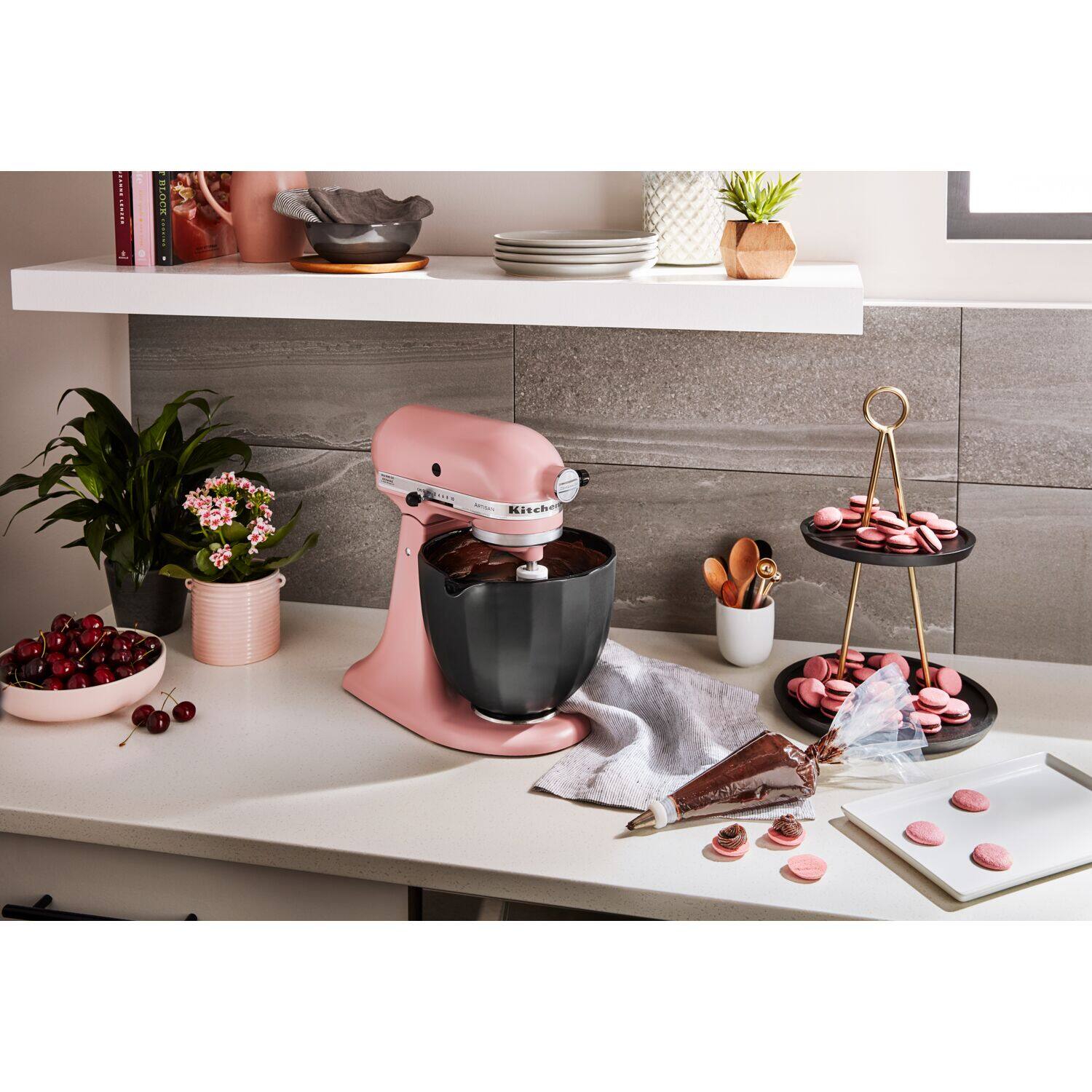 The image shows a kitchen counter with a pink food processor and a bowl of cookies. There are also some books on the counter, and a potted plant nearby. The scene is set against a wall with a shelf above it.