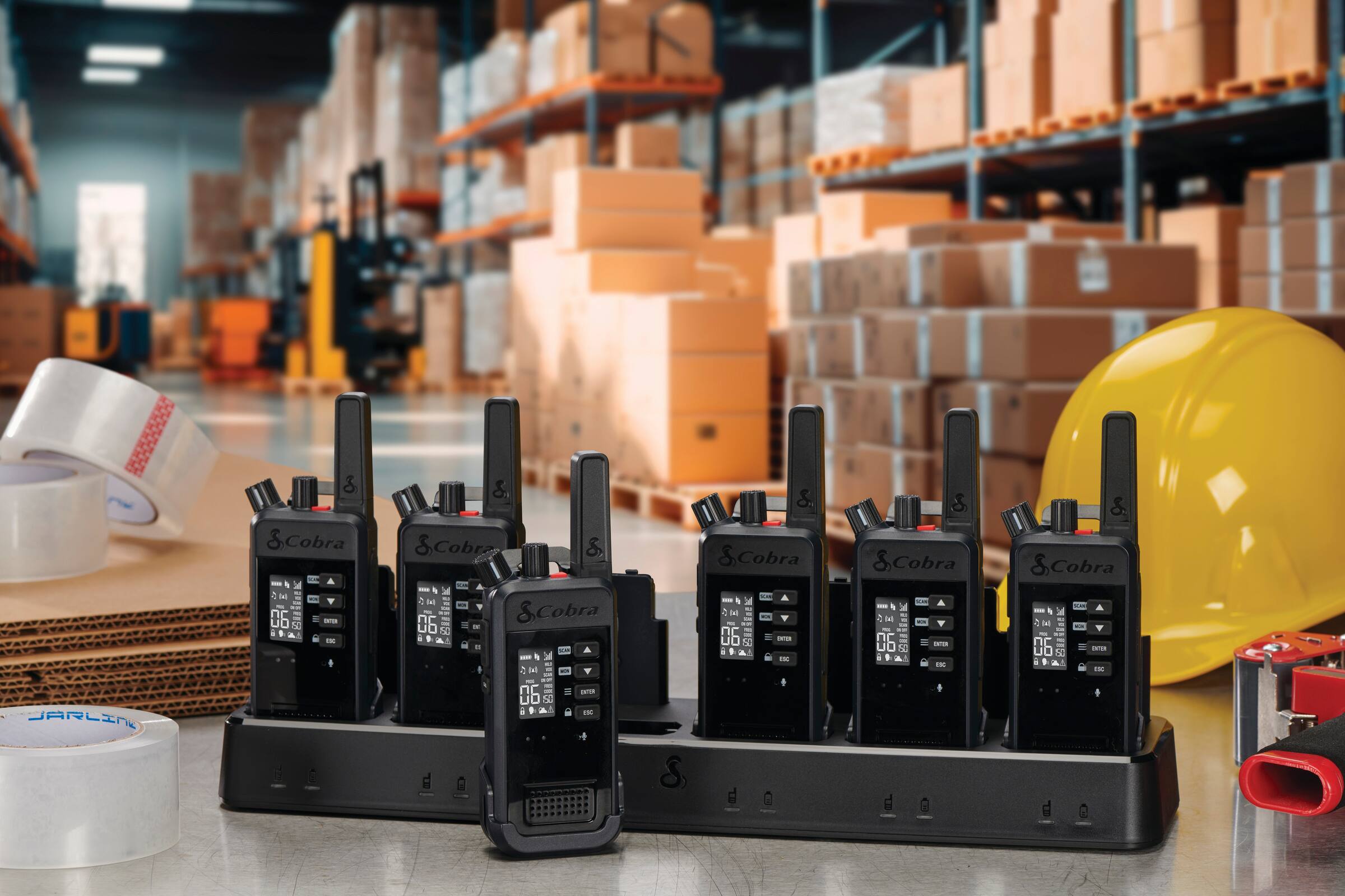 The image features a warehouse with a large number of boxes and pallets. There are also several radio devices, including walkie-talkies, placed on a table. The radio devices are labeled with barcodes, indicating that they are likely part of a larger inventory management system. The presence of the radio devices and barcodes suggests that the warehouse may be using these devices for communication and organization purposes.