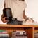 The image features a woman holding a black appliance, possibly a coffee maker, while standing next to a cat. The cat is sitting on a bookshelf, surrounded by several books. The books on the shelf include titles such as "Petlibro," "Mespe," "Lanpinza," "Cers Orgy," "The Hand That Feeds You," and "The Poetry of Pablo Neruda." The woman appears to be petting the cat, creating a cozy and relaxed atmosphere in the scene.