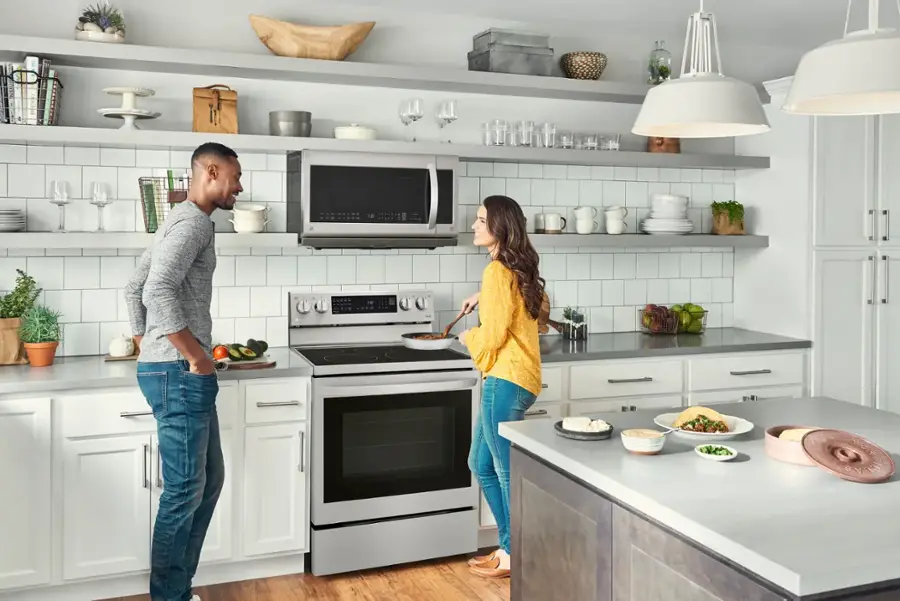 The image shows a man and a woman standing in a kitchen. The man is wearing a grey shirt, while the woman is wearing a yellow shirt. They are both standing in front of a stove, which is located under a microwave. The kitchen is well-equipped with various items such as a sink, a refrigerator, and multiple bowls and cups placed on the countertops. There is also a potted plant in the room, adding a touch of greenery to the space.