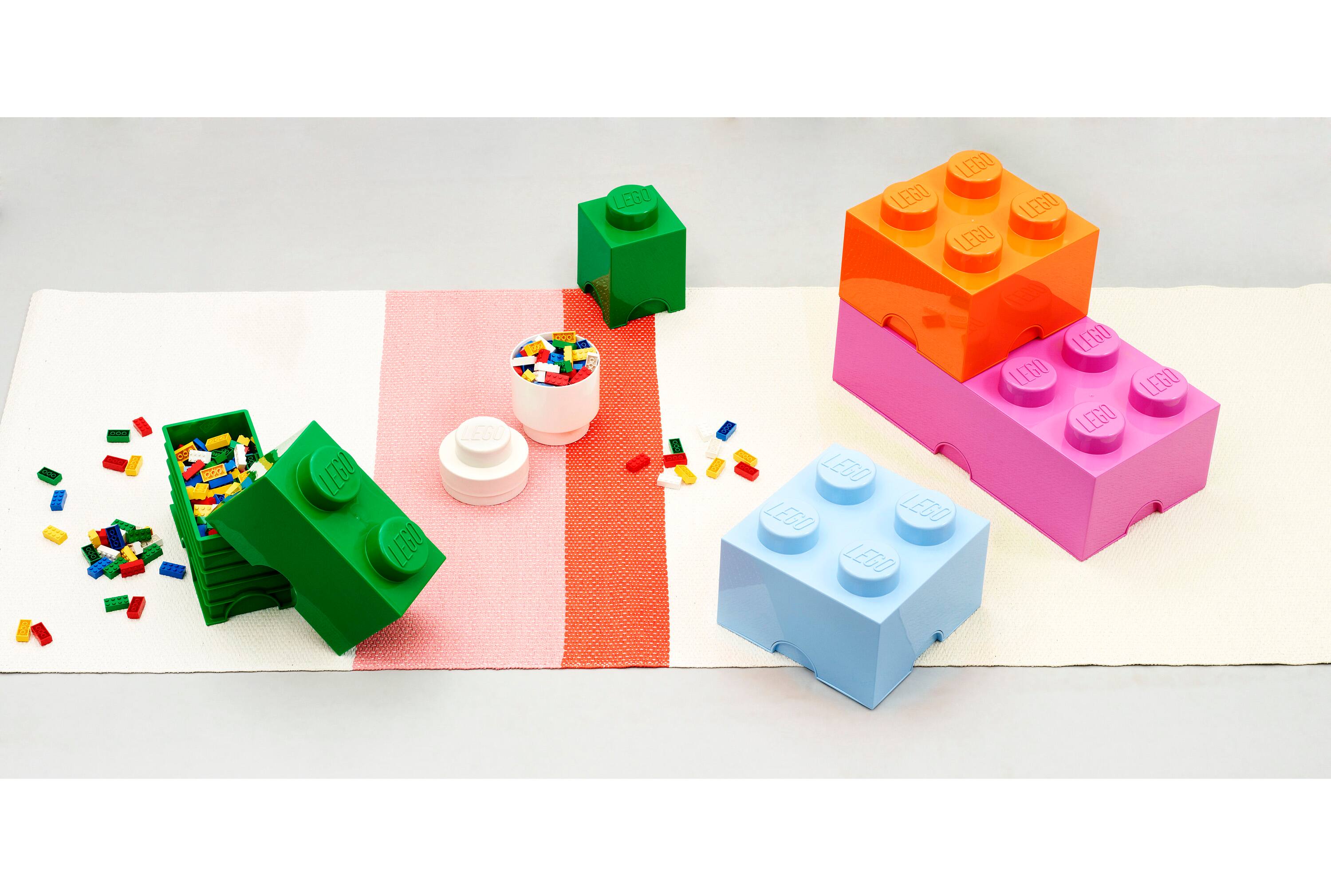 The image features a table with various Lego pieces, including a green Lego piece and a pink Lego piece. There are also several cups and bowls on the table, with one cup placed near the green Lego piece and two bowls located closer to the pink Lego piece. The table is covered with a white sheet, and the Lego pieces are arranged in a way that they appear to be building a house.