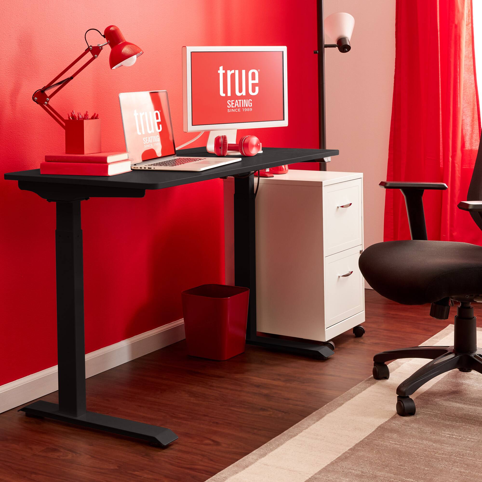The image features a red office desk with a laptop, a monitor, and a keyboard. There is also a red chair positioned in front of the desk. The desk is situated in a corner of a room with a red wall, and there is a red curtain in the background. The desk is equipped with a lamp, and there is a book placed on the desk. The overall scene suggests a workspace with a focus on productivity and comfort.
