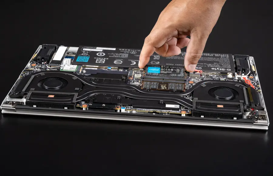 The image shows a person's hand pointing at a laptop computer, which is open and placed on a table. The laptop is equipped with a cooling fan, and it appears to be a MacBook Pro. The hand is pressing on the laptop's keyboard, possibly to demonstrate how to use the device or to showcase its features. The laptop's components, such as the cooling fan, are visible and highlighted in the image.