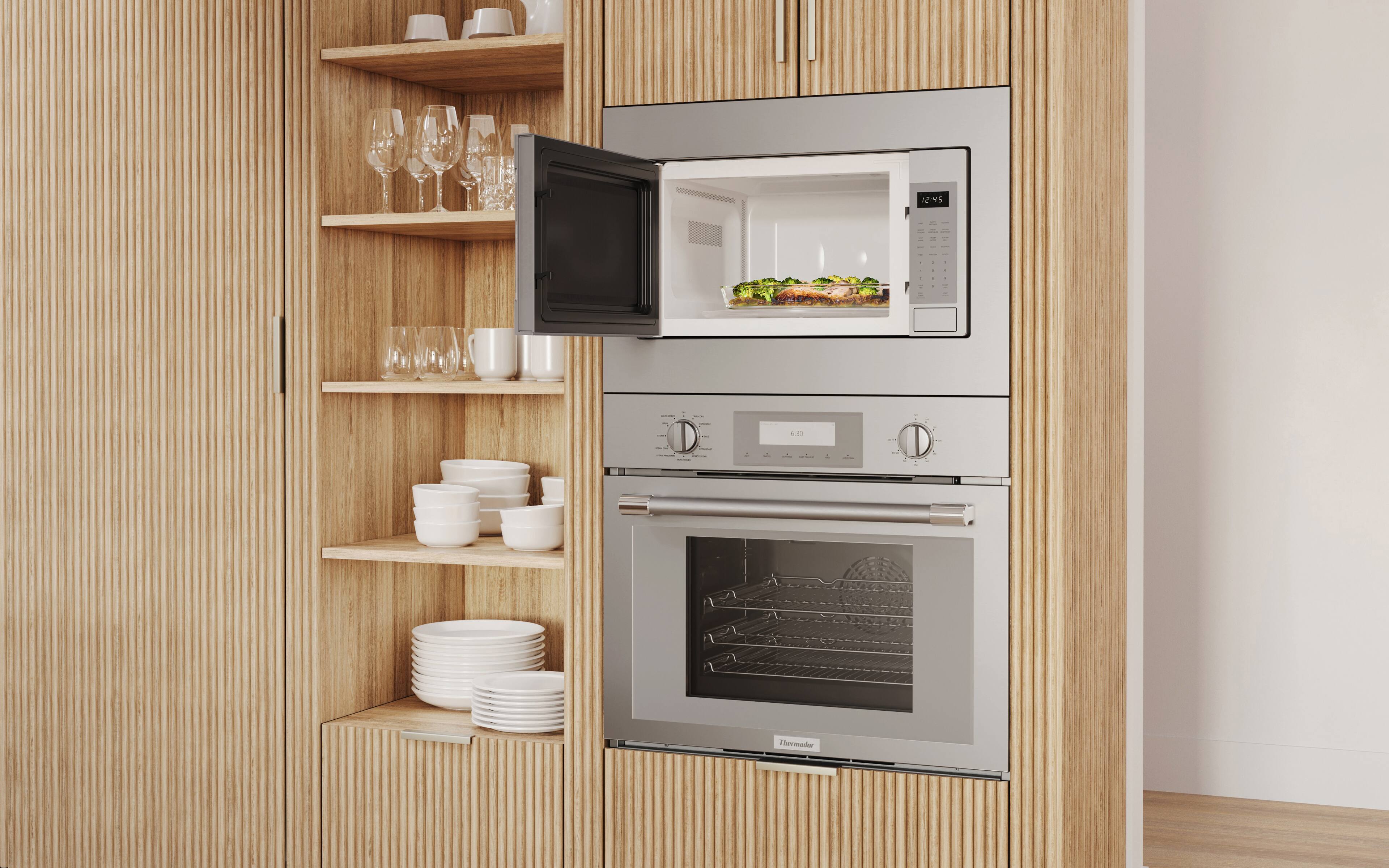 The image features a kitchen with a microwave oven built into a cabinet. The microwave is open, revealing a meal inside. The kitchen also has a stove top oven, and various items such as wine glasses, cups, and bowls are placed on the countertops. There are also several bottles and a sandwich visible in the scene. The image is described as having a "modern look" and "a lot of stuff."