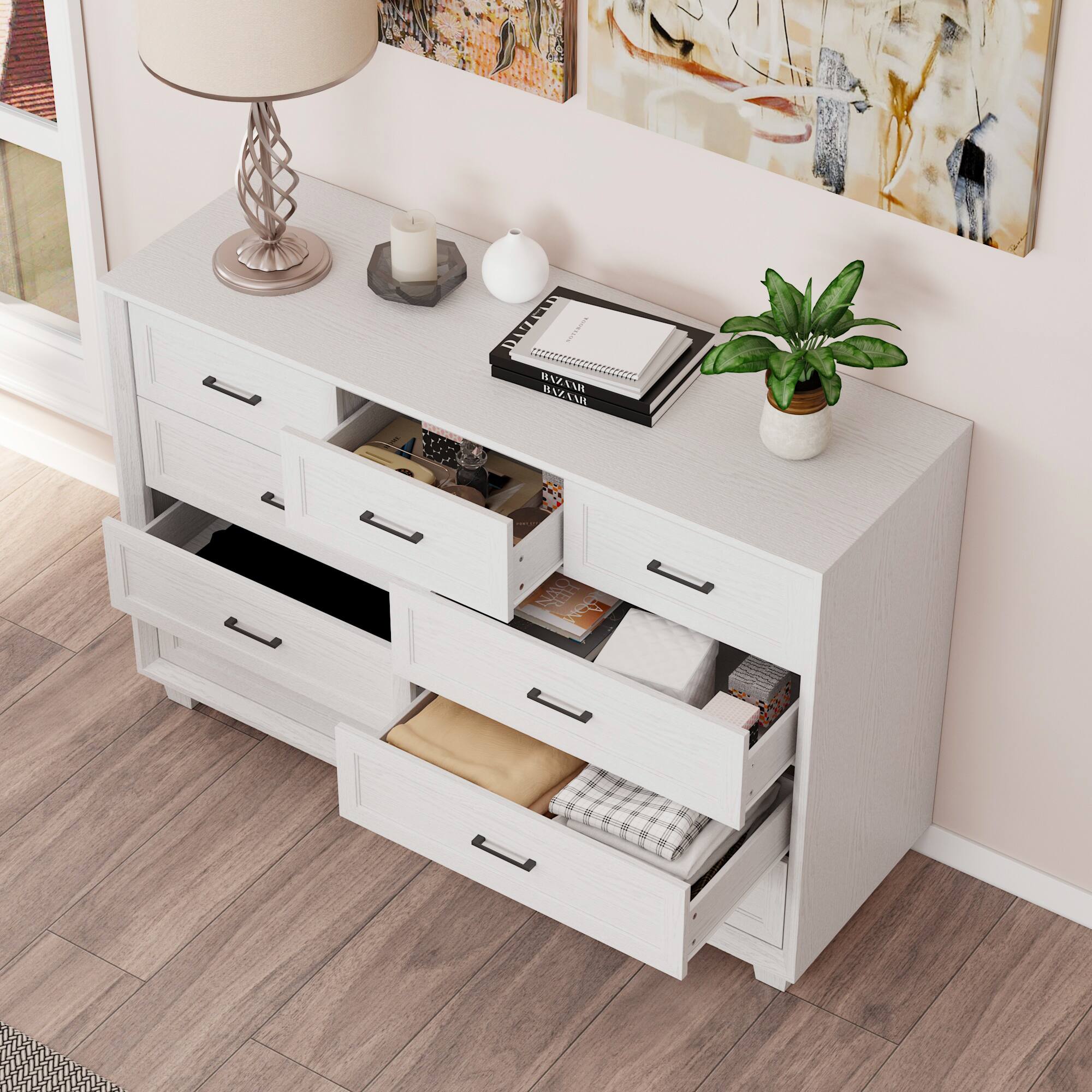 The image shows a white dresser with a variety of items on top of it. There is a lamp, a potted plant, and a book placed on the dresser. The dresser also has a drawer with a towel in it. The scene is set against a pink wall, and there is a window visible in the background.