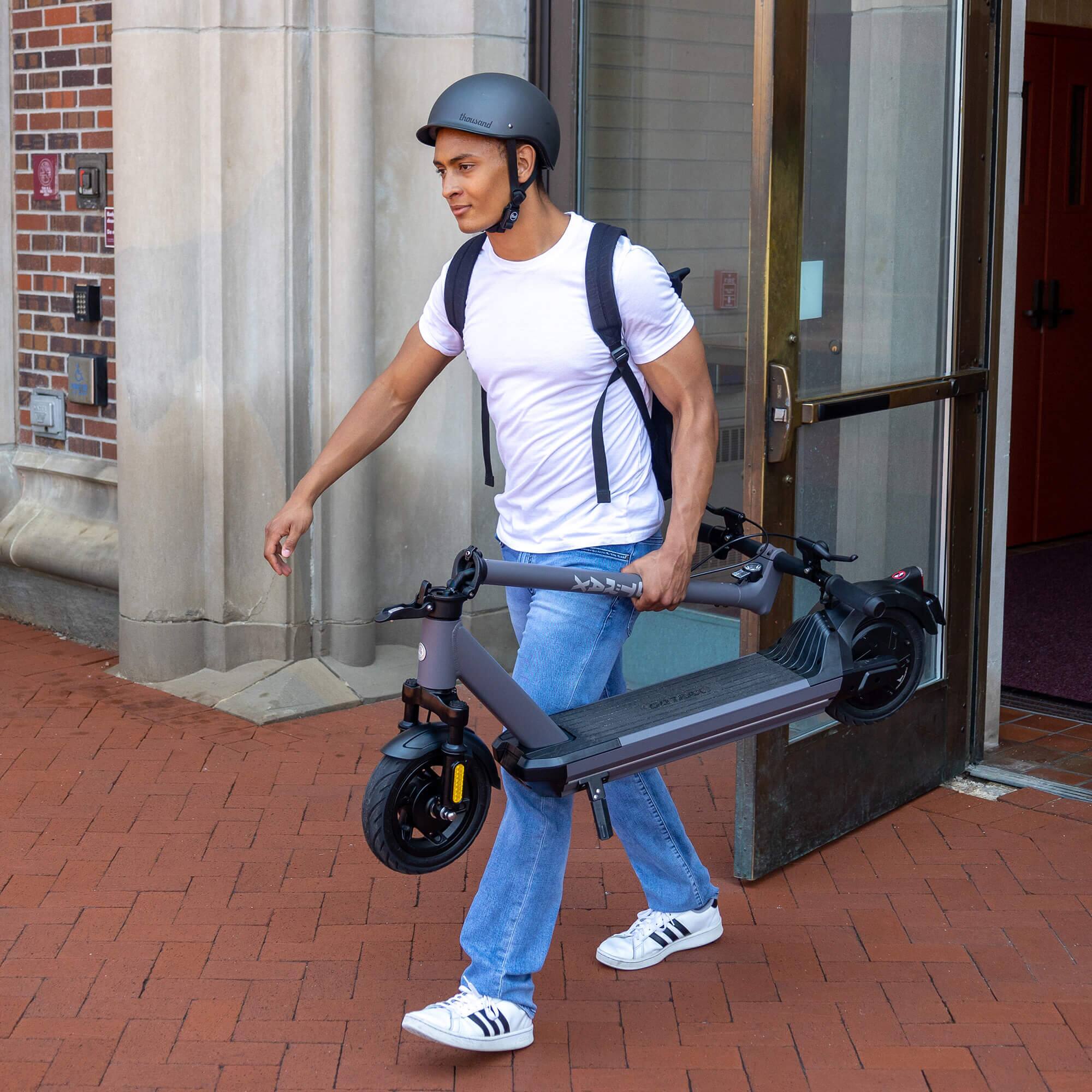 The man is wearing a helmet and carrying a scooter or a bike down the sidewalk.