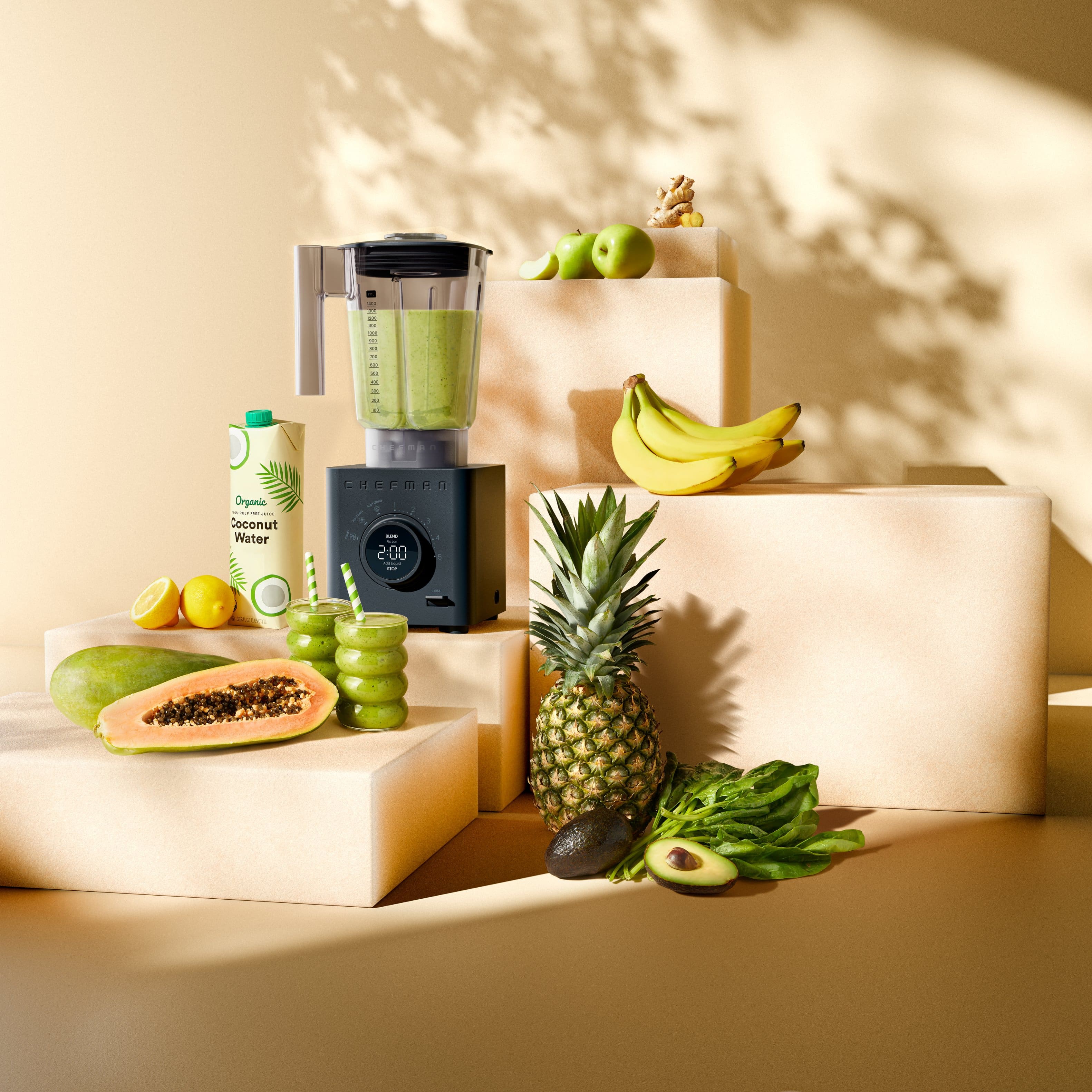 The image features a variety of fruits and a blender on a table. There are bananas, apples, and a pineapple, along with a coconut and a lemon. The blender is filled with green liquid, likely a smoothie or juice. The fruits and blender are arranged in a visually appealing manner, creating an inviting and healthy display.