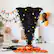 Halloween decorations are displayed in the living room. There is a black tree with orange and purple decorations, including a witch's hat. The room also features a pumpkin, a spider web, and a basket of oranges. The decorations create a festive atmosphere for the holiday.