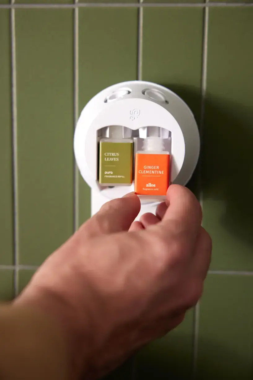 The image shows a person holding two small bottles of refill fragrance in their hand, with a dispenser on the wall. The bottles are labeled with the fragrances "Citrus Leaves" and "Ginger Clementine."
