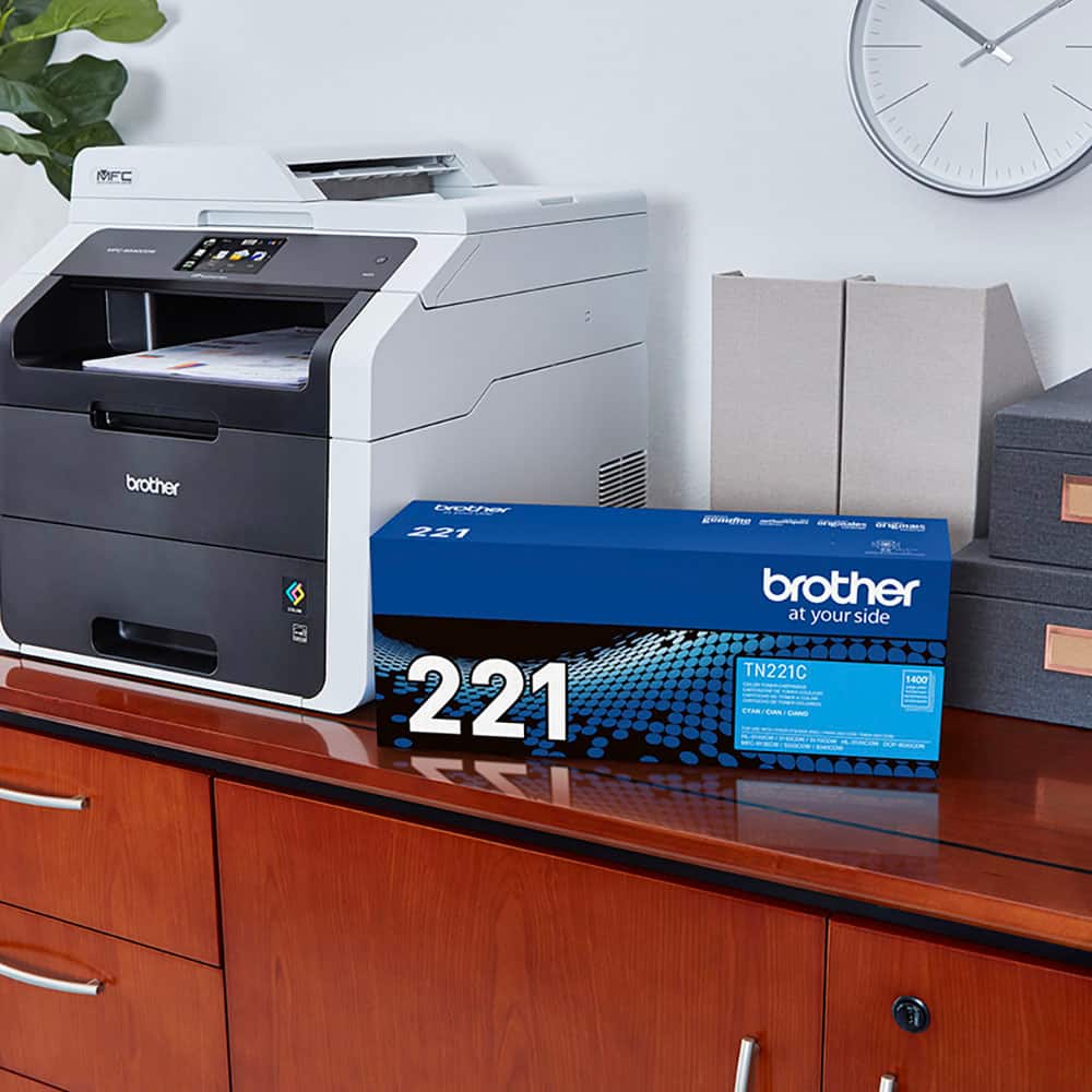 The image features a printer and a box of toner cartridges for the printer. The printer is a Brother MFC-221, and the toner cartridges are labeled "Brother at your side." The box of toner cartridges is placed next to the printer on a desk.