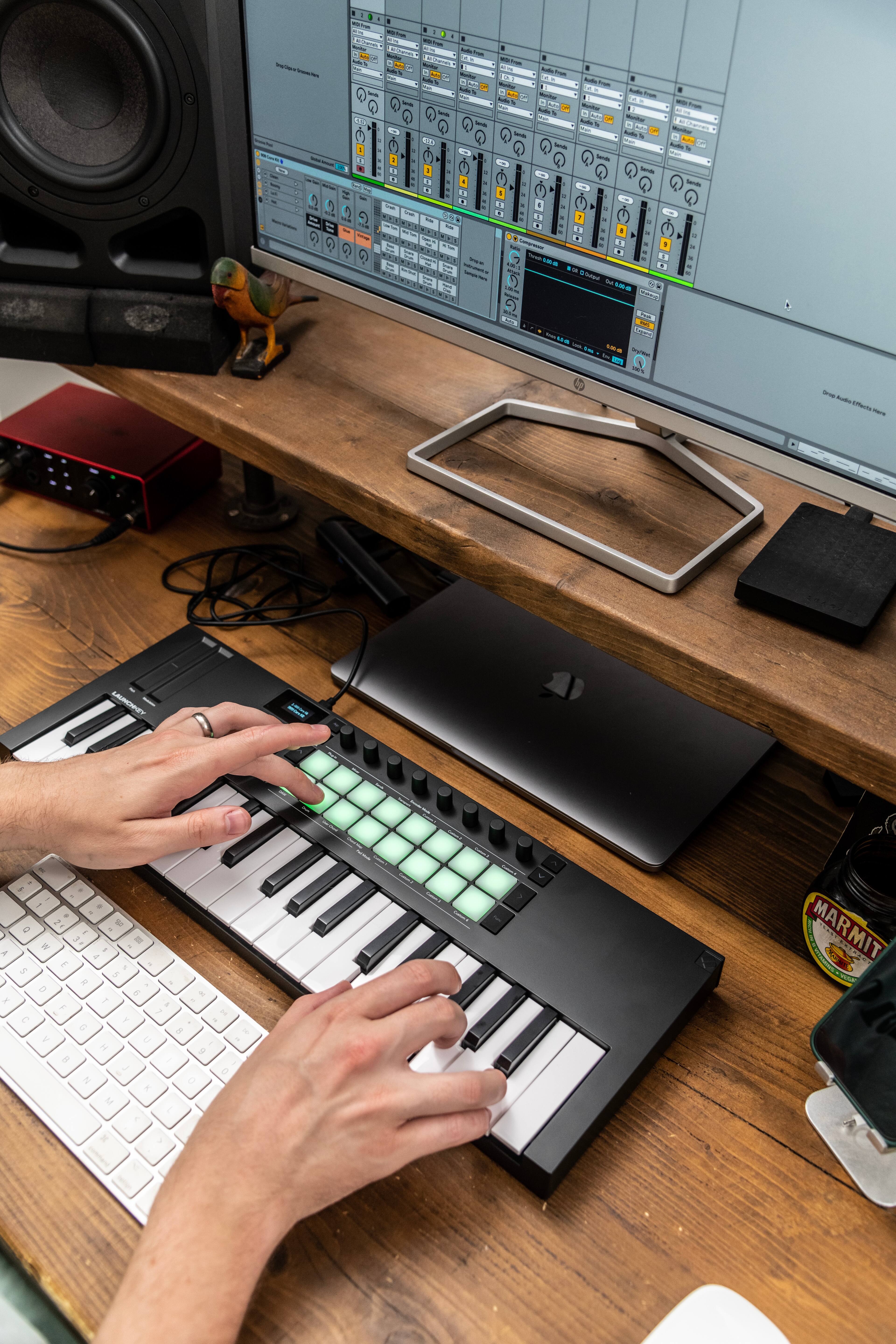 The image shows a person using a keyboard and a music keyboard, likely creating music or working on a project. The person is sitting at a desk with a computer monitor and a laptop. There are also two keyboards in the scene, one of which is a music keyboard. Additionally, there are two cups and a bottle on the desk, suggesting that the person might be enjoying a beverage while working.