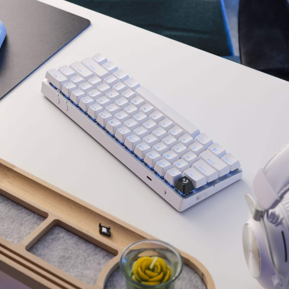 The image shows a white keyboard sitting on a white desk. The keyboard is placed in front of a black mouse pad. There is also a cup and a vase on the desk, adding to the overall aesthetic of the workspace.