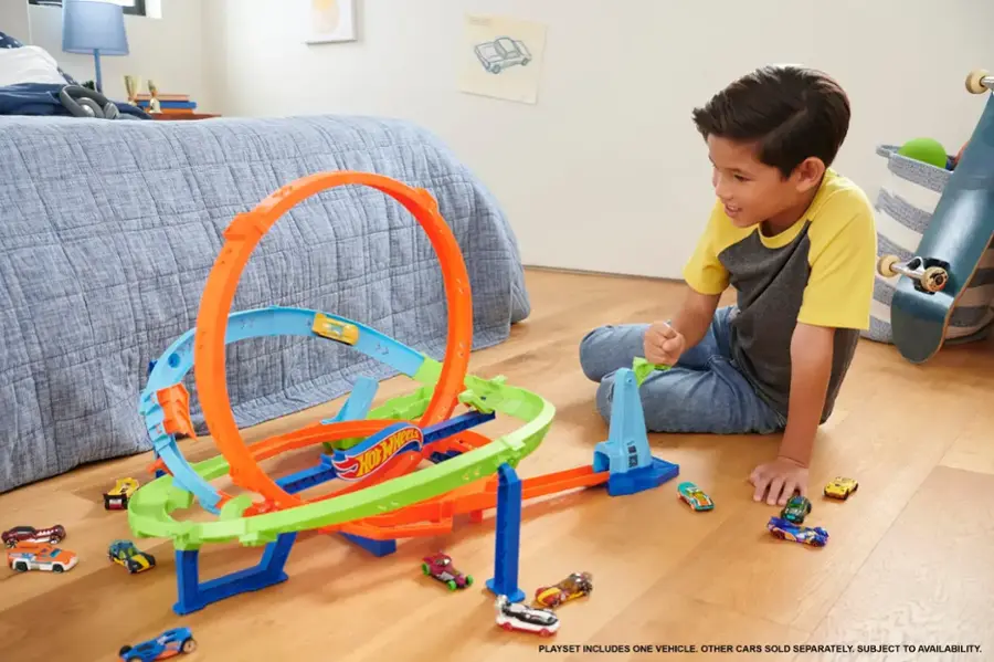 The image shows a young boy playing with a toy train set on a hardwood floor. The train set includes one vehicle, and other cars are sold separately. The availability of the toy cars is subject to availability.