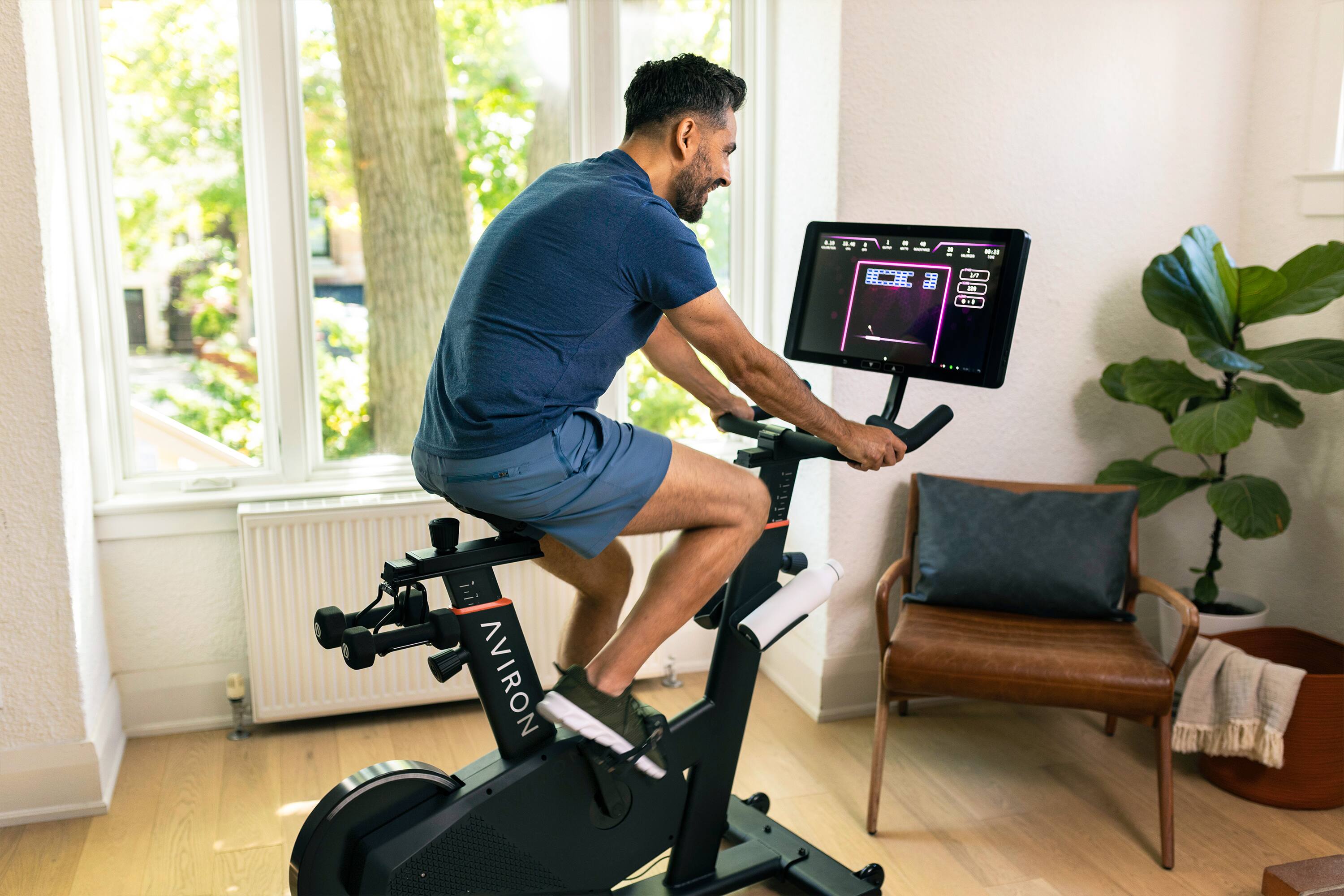 The man is riding a stationary bike indoors, likely in a living room. He is wearing a blue shirt and shorts, and there is a TV in front of him. The TV is displaying a video game, possibly a racing game, as the man appears to be playing it while riding the bike. The room also features a potted plant and a chair, creating a cozy and comfortable atmosphere for the man's indoor exercise session.