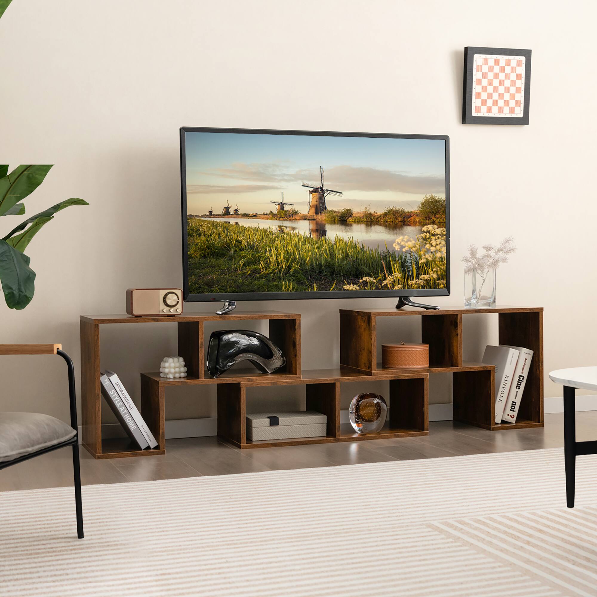 The image shows a living room with a television on a wooden stand. The television is displaying a picture of a windmill. The room also has a chair, a table, and a potted plant. There are several books scattered around the room, and a vase is placed on the table. The overall style of the room is modern and comfortable, with a touch of rustic charm from the wooden stand and the potted plant.