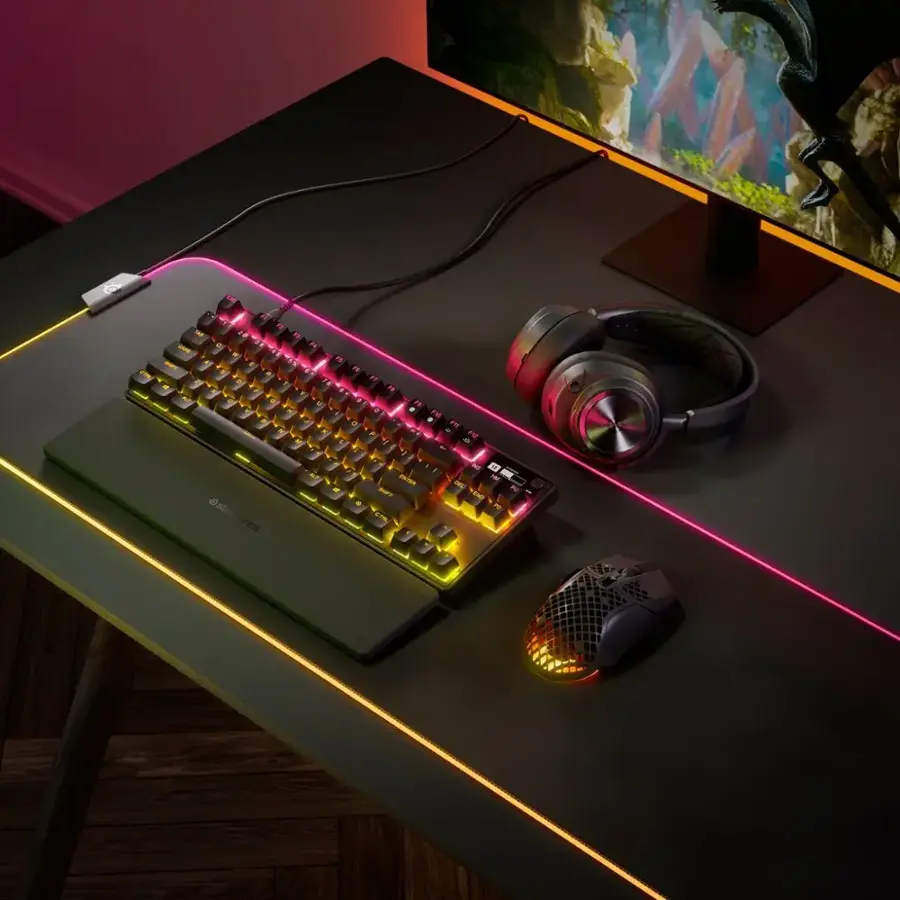 The image features a computer setup on a desk, including a keyboard, mouse, and headphones. The keyboard is a colorful, illuminated keyboard with a pink and yellow strip. The mouse is placed next to the keyboard, and the headphones are located nearby. The desk is positioned in front of a TV, which is turned on and displaying content. The overall scene suggests a comfortable and well-equipped workspace for various tasks and entertainment.