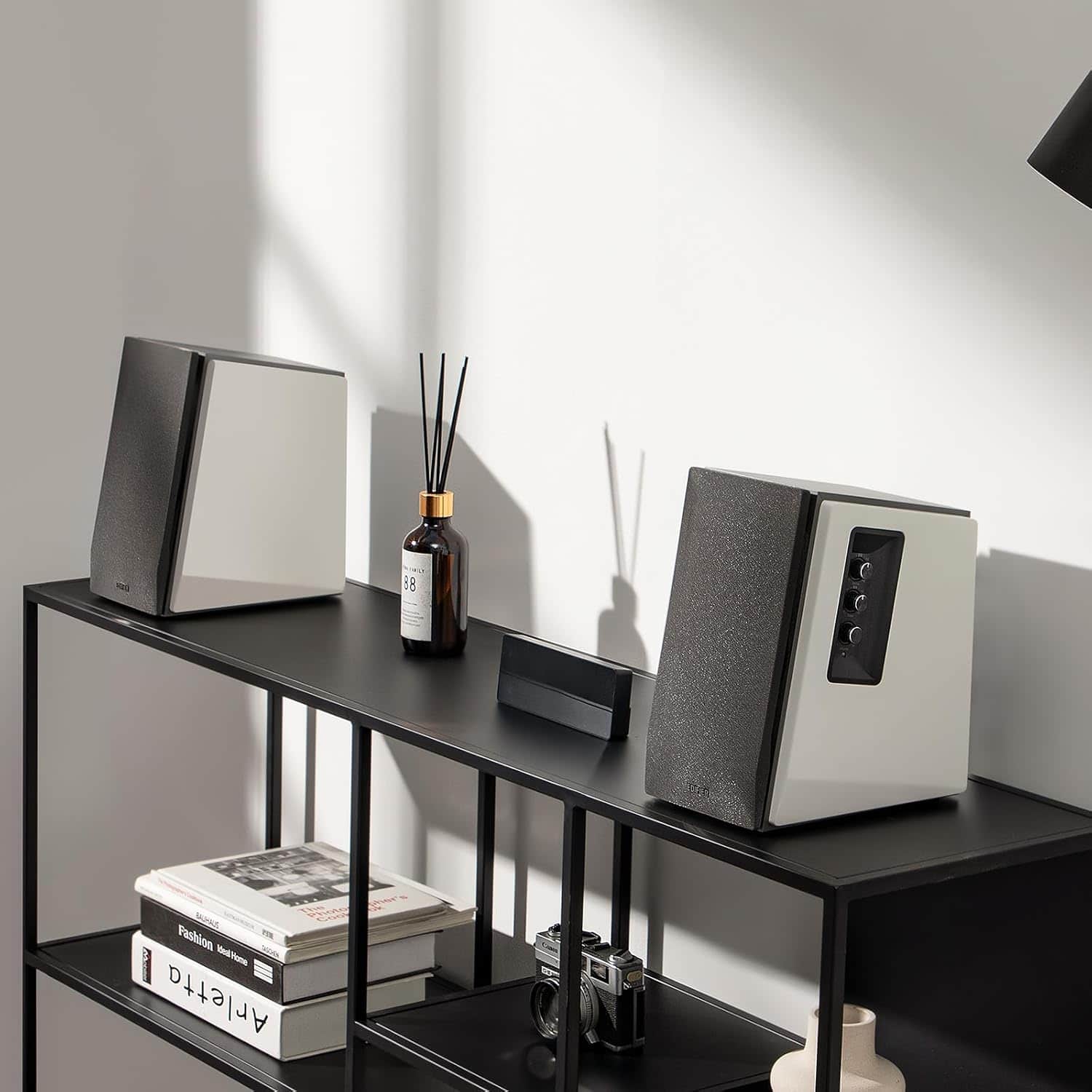 The image features a black table with two speakers on it. There is also a book on the table, and a bottle of oil is placed nearby. The table is situated in front of a wall, and there is a window in the background. The scene appears to be a modern and stylish living space.