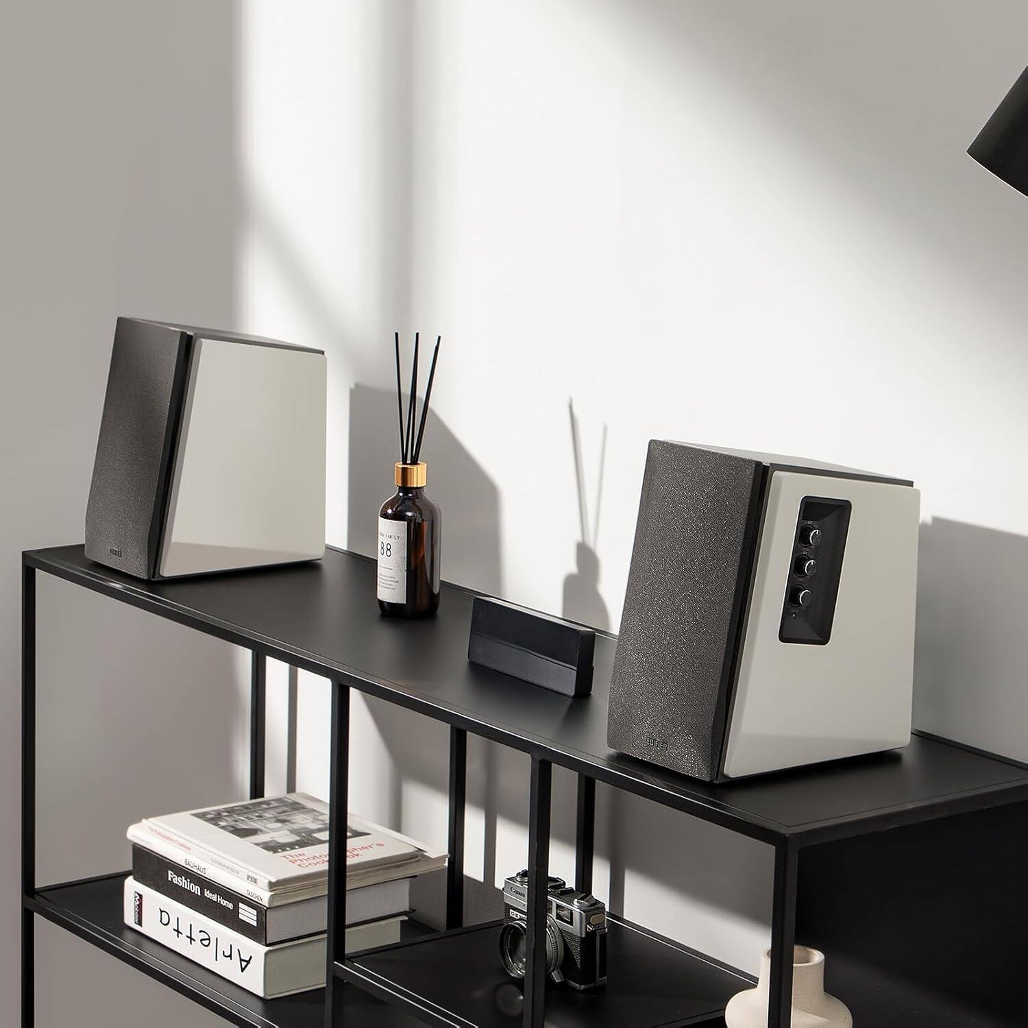 The image features a black table with two speakers on it. There is also a book on the table, and a bottle of oil is placed nearby. The table is situated in front of a wall, and there is a window in the background. The scene appears to be a modern and stylish living space.