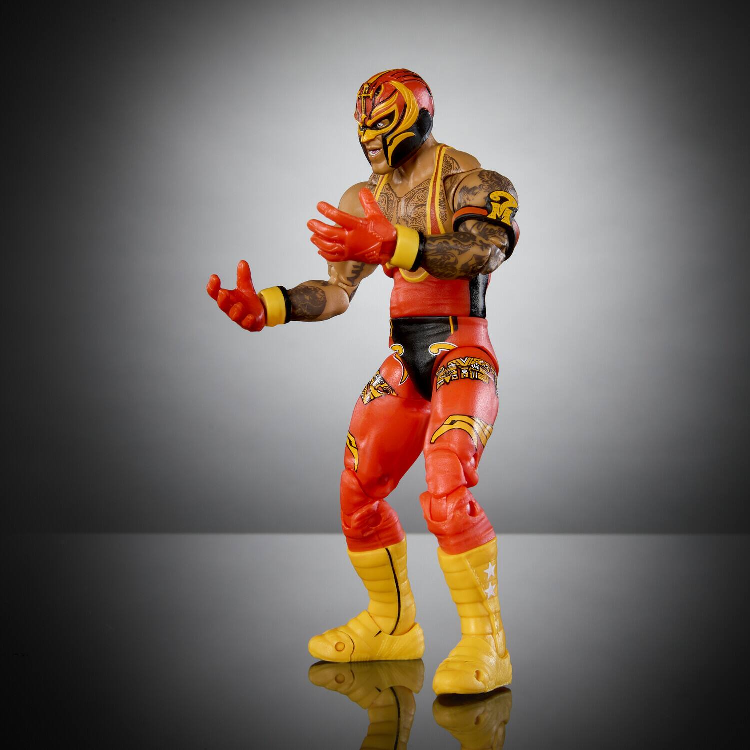 The image shows a wrestling action figure of a wrestler wearing a red and black mask, red and black vest, red and black pants, and yellow boots. The figure is posed with its hands outstretched.