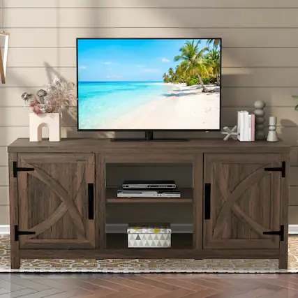 The image shows a wooden TV stand with a large flat-screen TV displaying a beach scene. The TV stand has two doors with X-shaped designs and a shelf with a DVD player and books. There are decorative items on the left, including a vase with dried flowers, and on the right, a stack of books and a small sculpture.