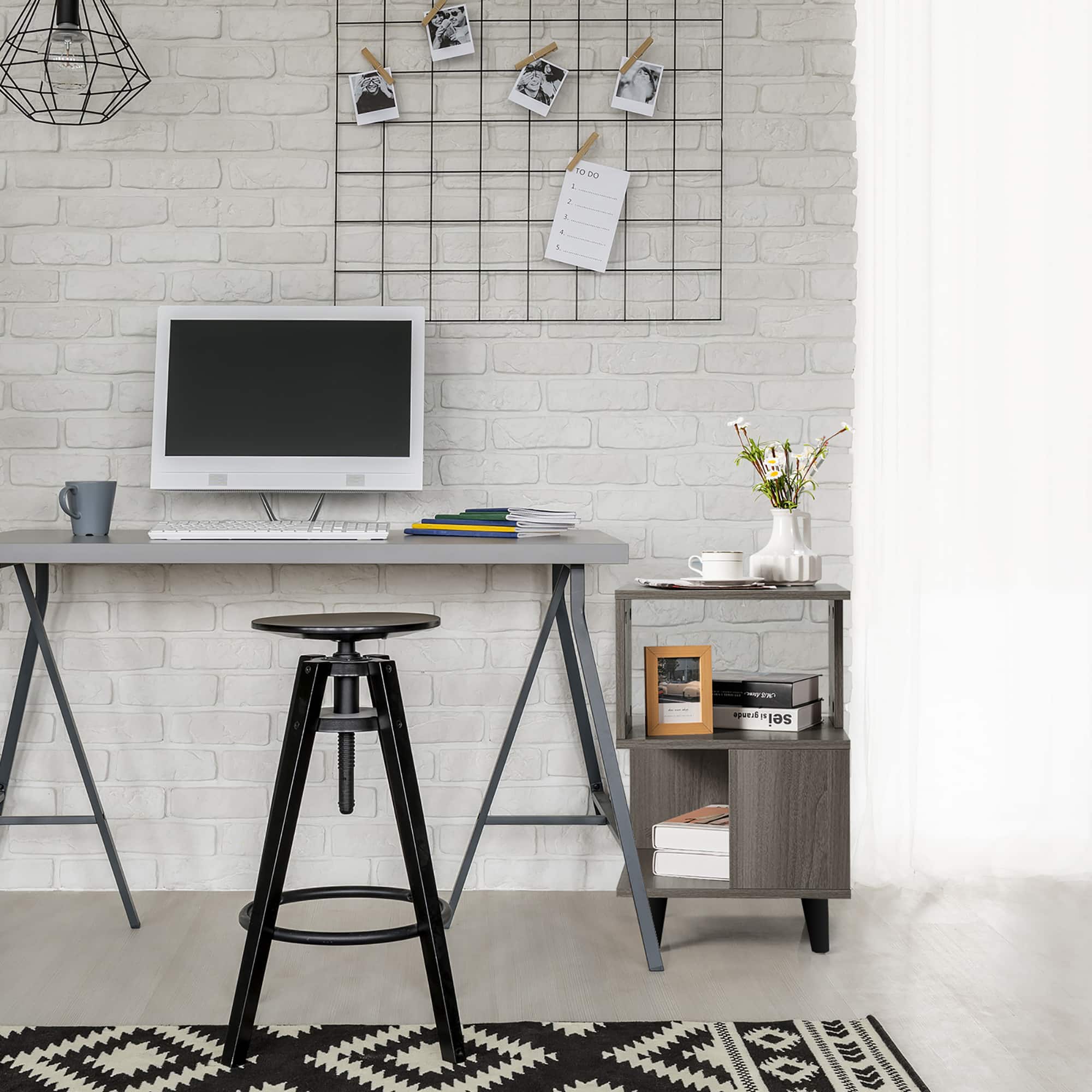 The image shows a small desk with a computer monitor, keyboard, and mouse on it. There is a chair placed in front of the desk, and a small table nearby. The desk is situated in a room with a brick wall, and there are several pictures hanging above the desk. Additionally, there is a vase with flowers on the desk, and a cup can be seen placed nearby. The overall setting appears to be a home office or workspace.