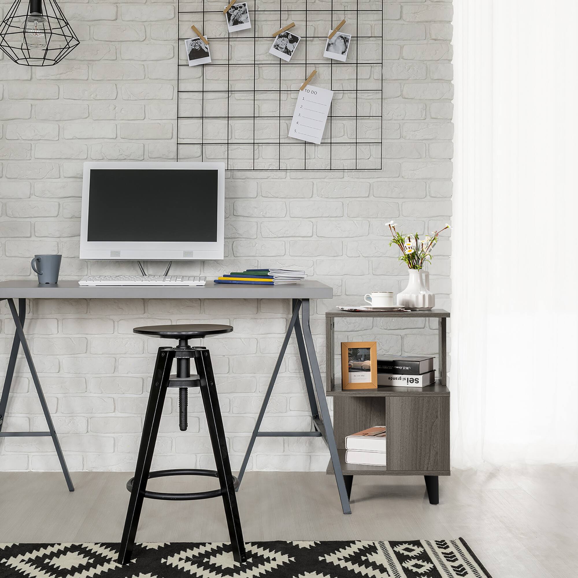 The image shows a small desk with a computer monitor, keyboard, and mouse on it. There is a chair placed in front of the desk, and a small table nearby. The desk is situated in a room with a brick wall, and there are several pictures hanging above the desk. Additionally, there is a vase with flowers on the desk, and a cup can be seen placed nearby. The overall setting appears to be a home office or workspace.