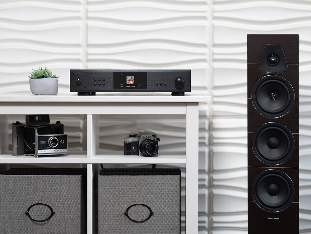 The image features a white desk with various electronic devices and equipment on it. There is a Nikon camera, a Polaroid camera, and a Rotel amplifier, all placed on the desk. Additionally, there is a speaker and a potted plant nearby. The desk is set up for a home entertainment system, with the electronic devices arranged to create an organized and functional space.