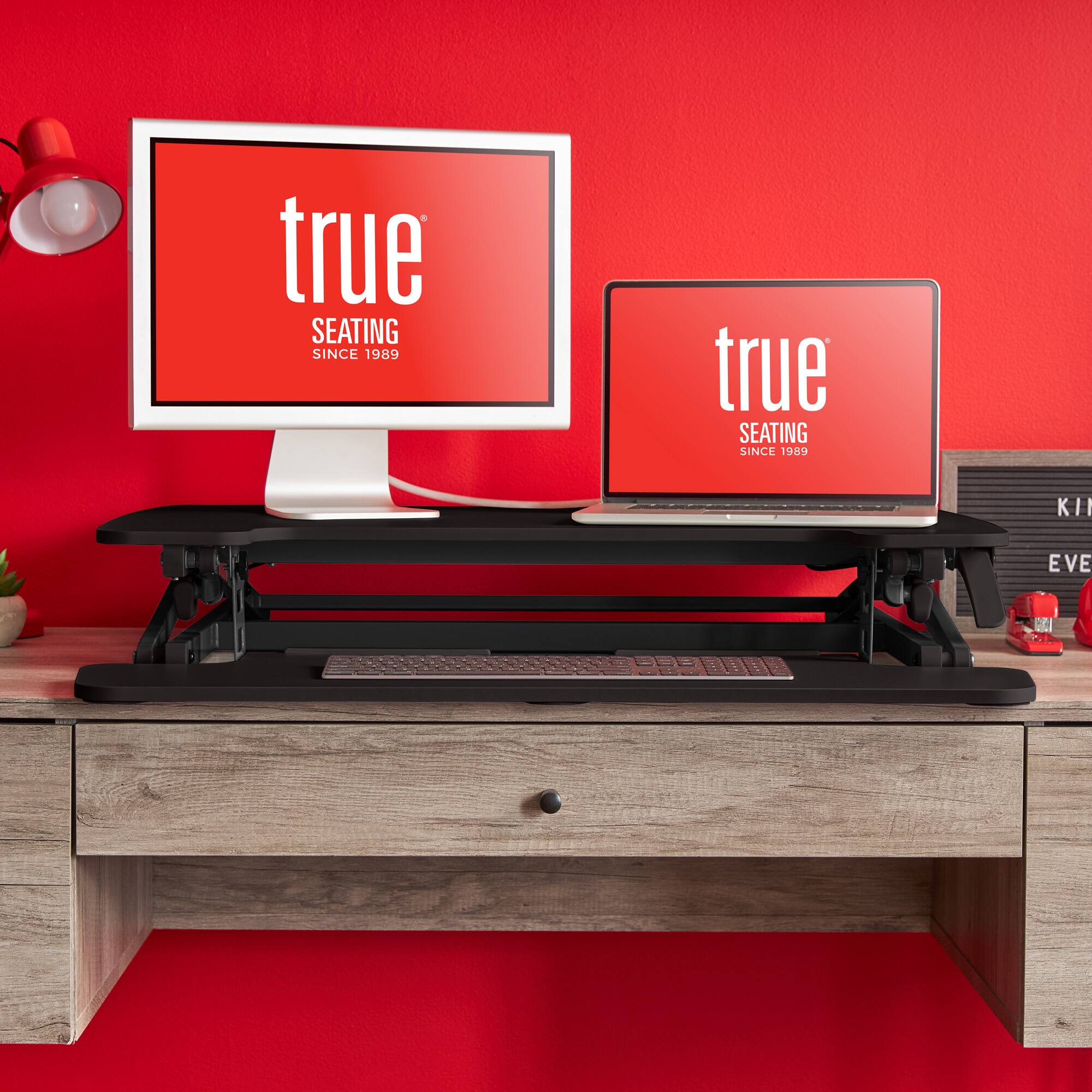 The image features a desk with a laptop and a computer monitor on it. The laptop is placed on a stand, and the computer monitor is positioned next to it. The desk is set against a red wall, and there is a potted plant nearby. The desk also has a keyboard and a mouse on it. The image is advertising a product called "True Seating," which has been in business since 1989.