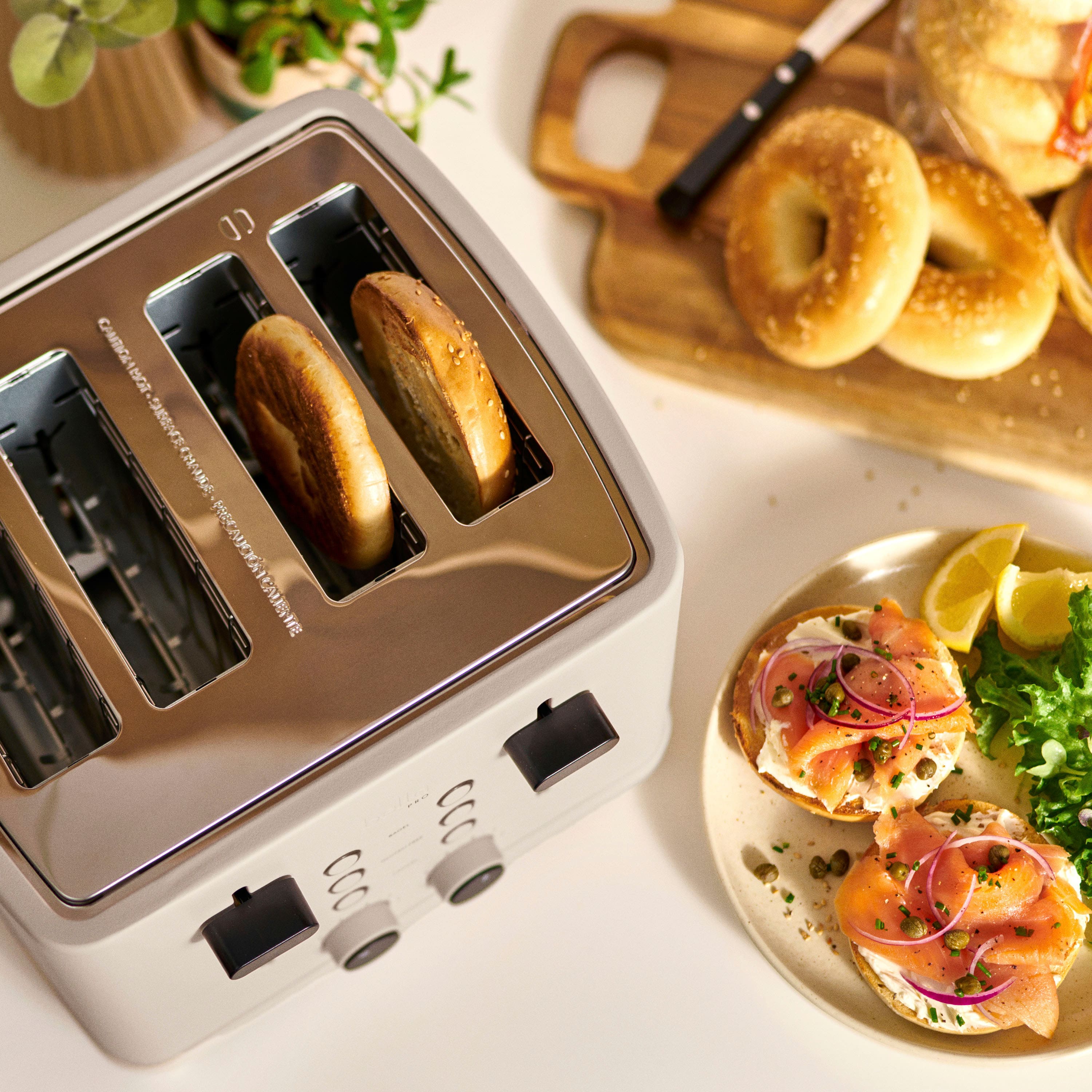 The image features a toaster oven with four bagels inside, placed on a counter. The toaster oven is made by Cuisinart and is filled with bagels. There is also a plate of food next to the toaster oven, which includes bagels and salmon. A knife is visible in the background, likely used for spreading or cutting the food. The scene suggests a breakfast or snack setting with a variety of food items available.
