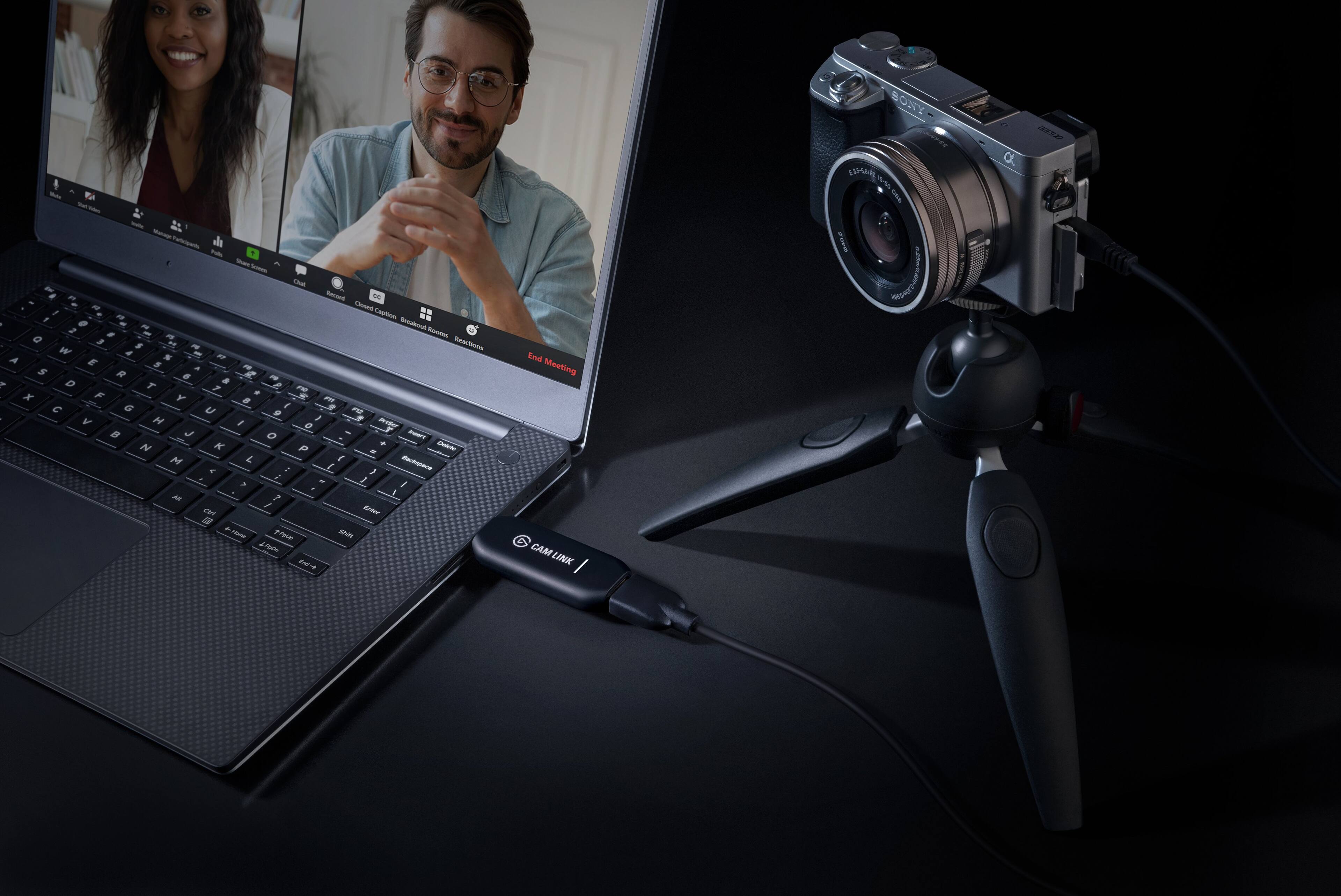 The image features a laptop computer with a webcam attached to it. The webcam is positioned in front of the laptop, likely for video conferencing or streaming purposes. The laptop is placed on a table, and there are two people visible in the image, one on the left side and the other on the right side. The scene suggests that the laptop is being used for a video call or a live broadcast, with the webcam capturing the participants' images and audio.