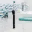 The image features a black and silver electric toothbrush standing upright on a white counter. The toothbrush is positioned in front of a sink, which is accompanied by a towel. The toothbrush is equipped with a green bristle head, making it suitable for daily oral hygiene routines.