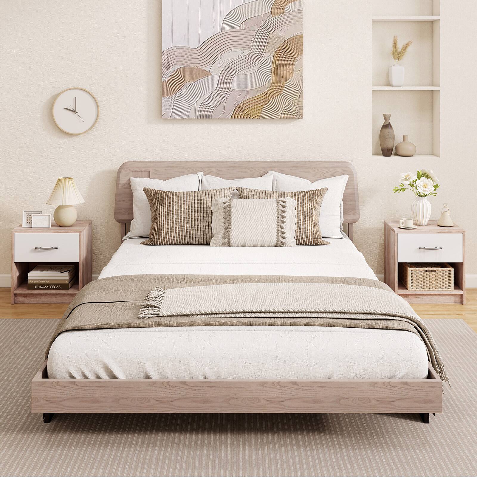 The image shows a neatly arranged bedroom with a wooden bed frame and a beige bedspread. The bed is adorned with multiple pillows and a folded blanket at the foot. On either side of the bed are matching wooden nightstands, each with a lamp, a book, and a small decorative item. Above the bed, there is a large abstract painting, and to the right, there are shelves with vases and a bouquet of flowers. A clock is mounted on the wall to the left. The room has a light, neutral color palette, creating a calm and cozy atmosphere.