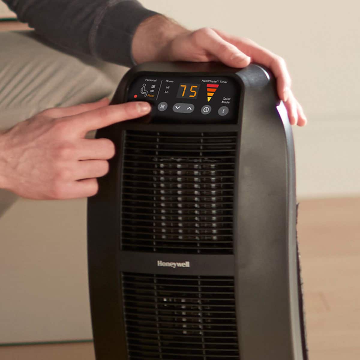 The image shows a person pointing at a Honeywell heater, which is placed on the floor. The heater has a digital display that shows the current temperature and mode. The person is likely demonstrating the heater's features or discussing its performance.