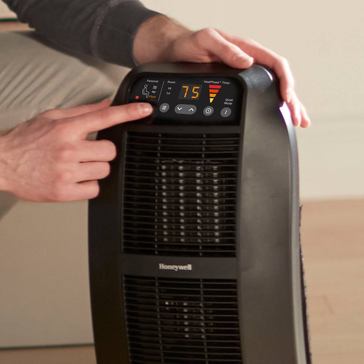 The image shows a person pointing at a Honeywell heater, which is placed on the floor. The heater has a digital display that shows the current temperature and mode. The person is likely demonstrating the heater's features or discussing its performance.