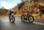 A man and a woman are riding bikes down a road. The man is wearing a red jacket and riding a bicycle with a child seat on the back. The woman is riding a bike behind him.