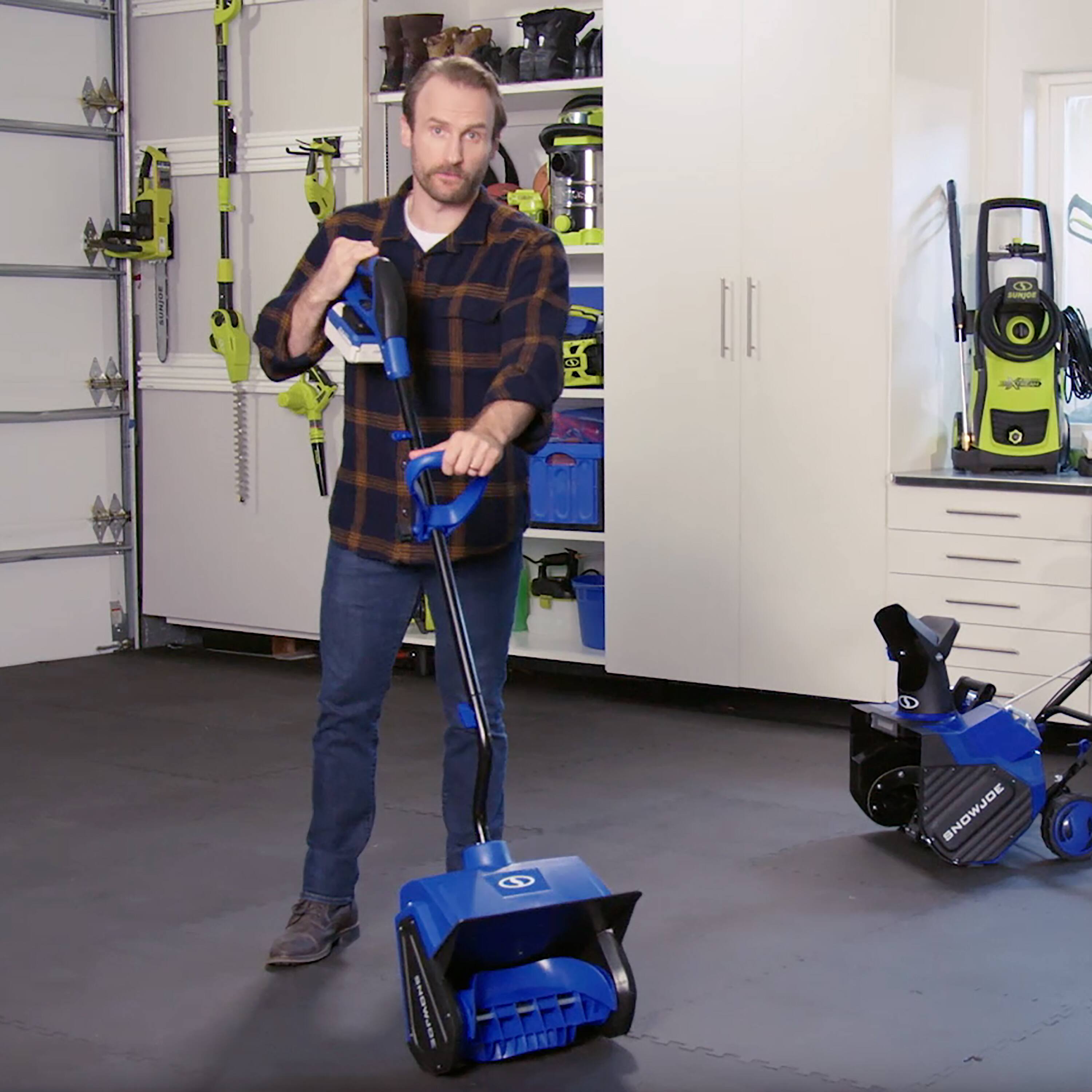 The man is standing in a garage with a blue snow blower and a blue lawn mower. He is holding a blue push broom, which is also blue in color. The garage is filled with various tools and equipment, including a pair of scissors and a handbag. The man appears to be in the process of cleaning or organizing the garage.