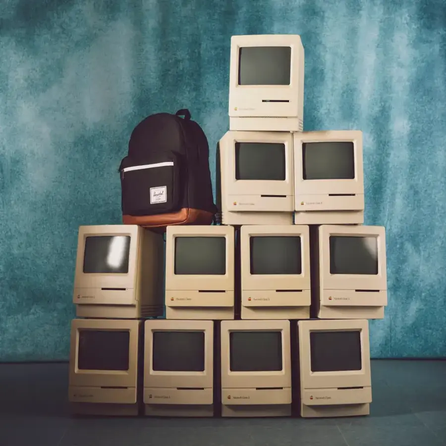 The image features a stack of old computer monitors and a backpack. The monitors are arranged in a pyramid shape, with some of them being stacked on top of each other. The backpack is placed on top of the stack of monitors, creating an interesting contrast between the old technology and the modern backpack.