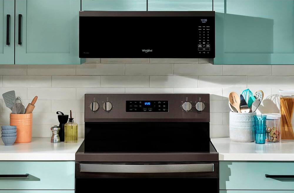 The image shows a kitchen with a black stove top oven. The oven is positioned under a microwave, which is placed above the stove. The kitchen counter is cluttered with various items, including a knife set, a spoon, a bowl, and a cup. The microwave is displaying the time 12:20. The kitchen is well-equipped and ready for cooking.