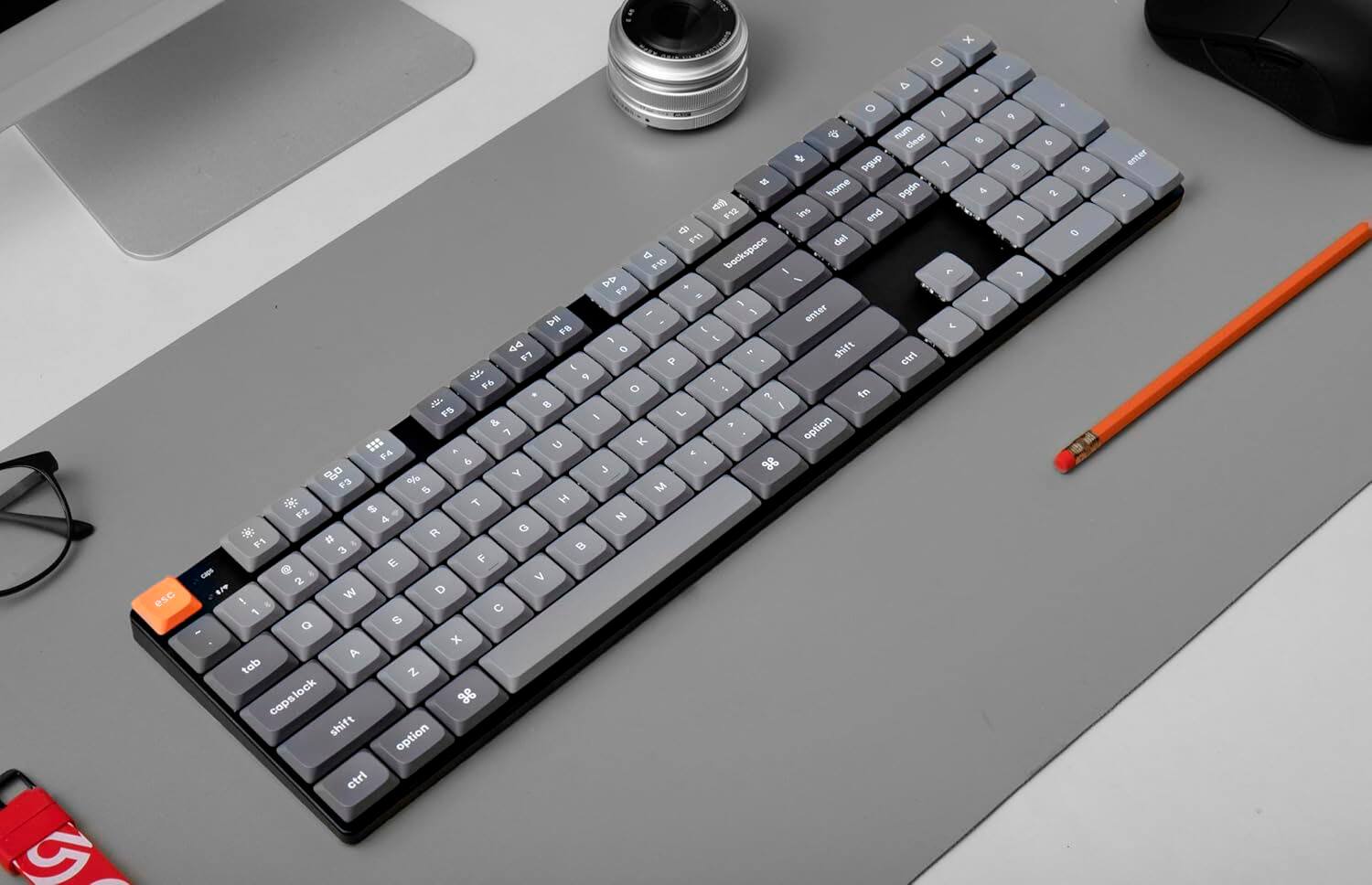 The keyboard is a compact keyboard with a grey and black color scheme. It has a combination of letters and numbers, including the numbers 1-9, the letters A-Z, and various function keys. The keyboard also features a few special keys, such as the escape key (Esc), the function keys F1-F12, and the arrow keys. Additionally, there is a space bar and a backspace key for easy typing. The keyboard is placed on a grey table, and there is a pencil nearby.