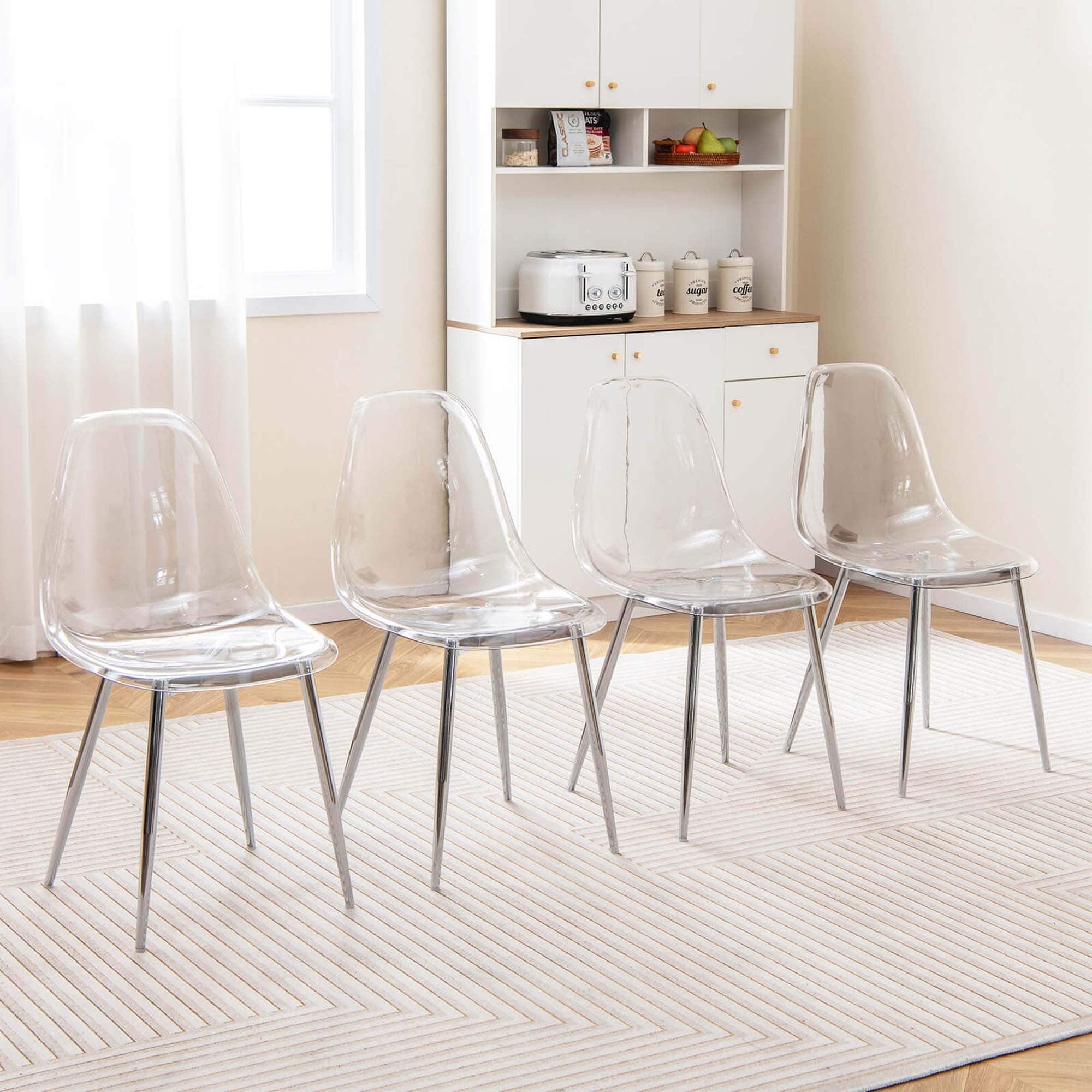 The image shows a living room with a white rug and four clear chairs arranged in a row. The chairs are positioned in front of a white cabinet, and there is a window nearby. The room also features a kitchen area with a sink and a bowl. Additionally, there are a few apples on the countertop.