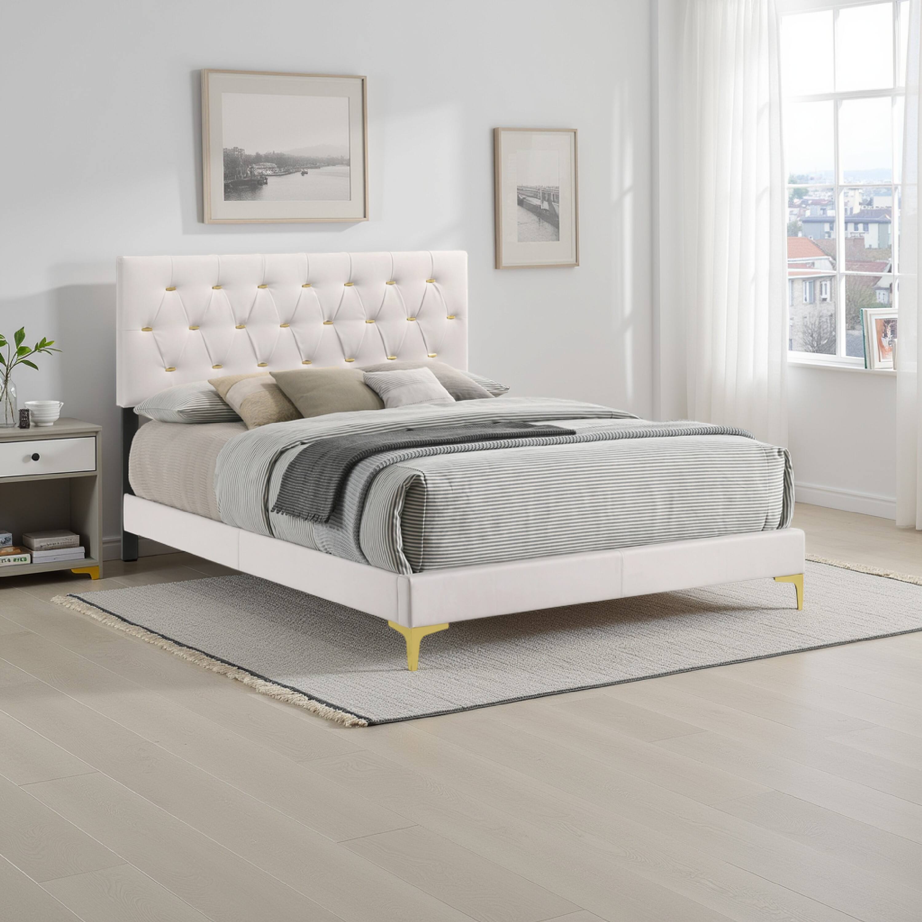 The image shows a modern bedroom with a white upholstered bed featuring a tufted headboard. The bed is dressed with a gray and white striped duvet and several pillows. On the left side, there is a nightstand with a small plant, a cup, and a few books. The room has light-colored wooden flooring and a light gray rug. Two framed pictures hang on the wall above the bed, and a large window with sheer curtains allows natural light to fill the room.