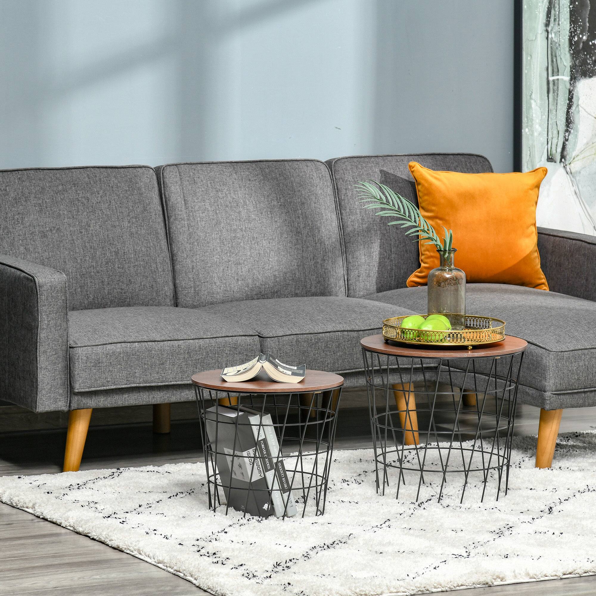 The image shows a modern living room setting featuring a grey fabric sofa with wooden legs. On the sofa, there is an orange cushion and a small vase with green foliage. In front of the sofa, there are two round coffee tables with a wireframe design. One table holds a book and a small box, while the other has a golden tray with green apples. The floor is covered with a white shaggy rug.