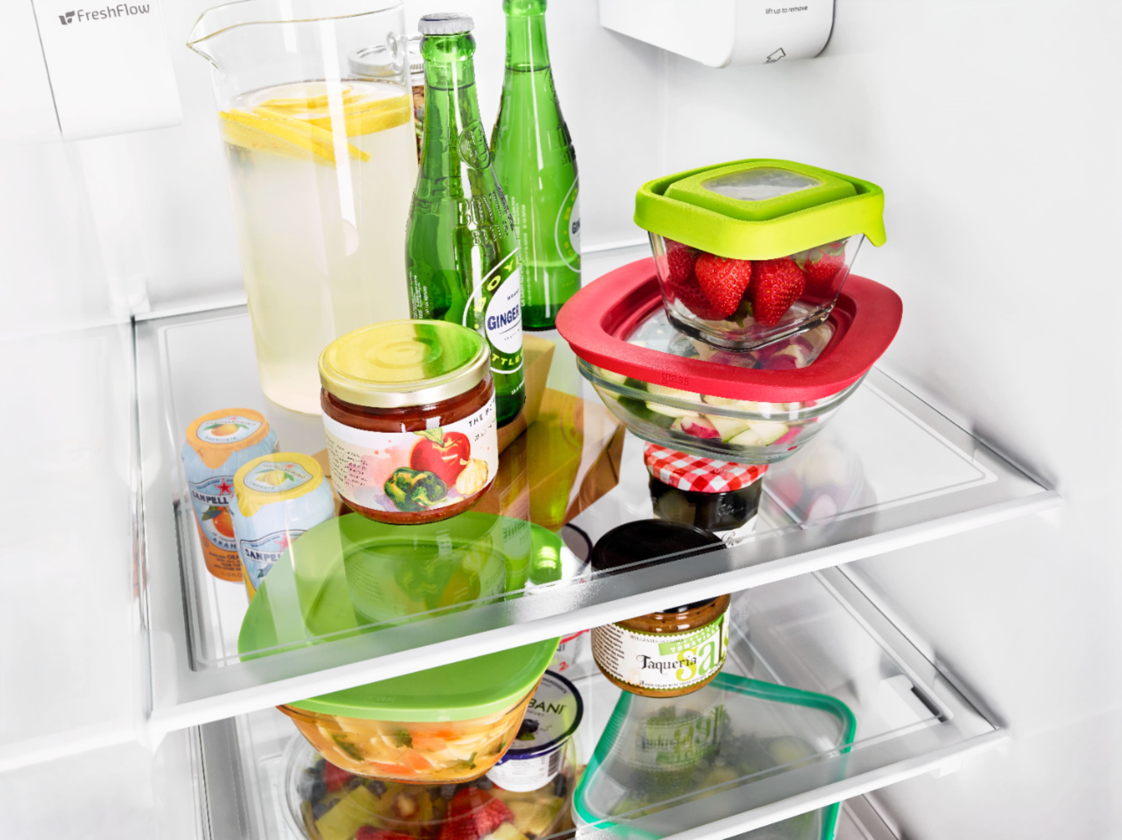 The image features a white refrigerator with a variety of food items and drinks inside. There are several bottles, including a green bottle, and a bowl placed on the shelves. The refrigerator is filled with food and drinks, showcasing a diverse selection of items.