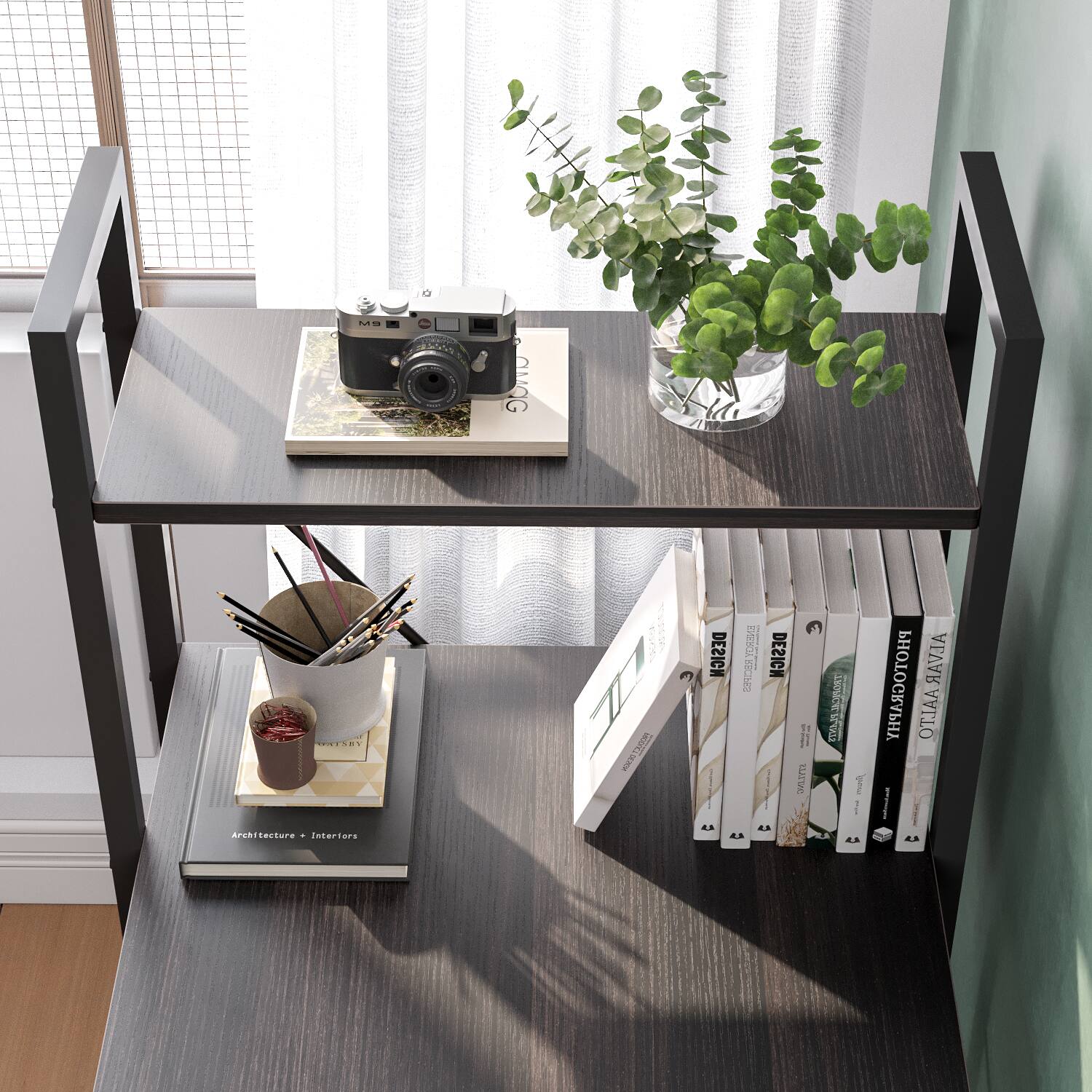 The image features a wooden desk with a variety of items on it. There is a bookshelf with several books, including a book about Alvar Aalto. A vase with a plant is also present on the desk, adding a touch of greenery to the space. Additionally, there is a camera and a cup on the desk, indicating that the person using the desk might be involved in photography or simply enjoys having a beverage nearby while working or studying. The overall scene suggests a well-organized and functional workspace.