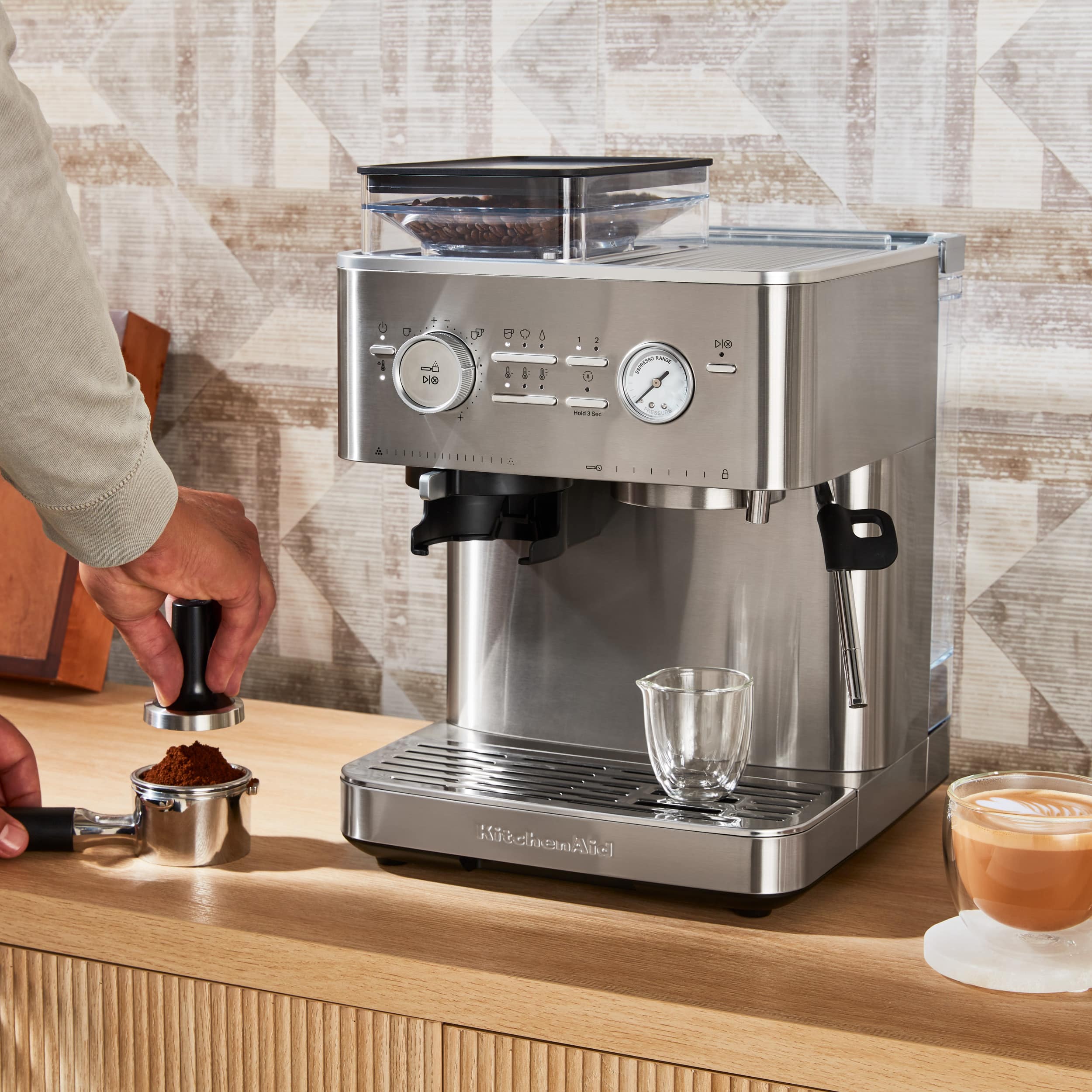 The image features a person using a coffee maker, which is placed on a counter. The coffee maker is equipped with a timer and a coffee cup, and it is ready to brew coffee. The person is in the process of preparing a cup of coffee, and there is a spoon nearby. The scene also includes a dining table and a chair, suggesting that the coffee maker is set up in a cozy and comfortable space for enjoying a hot beverage.
