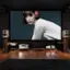 The image features a woman wearing headphones and a white shirt, standing in front of a large screen. The screen displays a picture of the woman, and there are several books scattered around the room. The room also has a couch, a speaker, and a TV. The woman appears to be posing for the picture, and the overall setting seems to be a living room or a multimedia room.